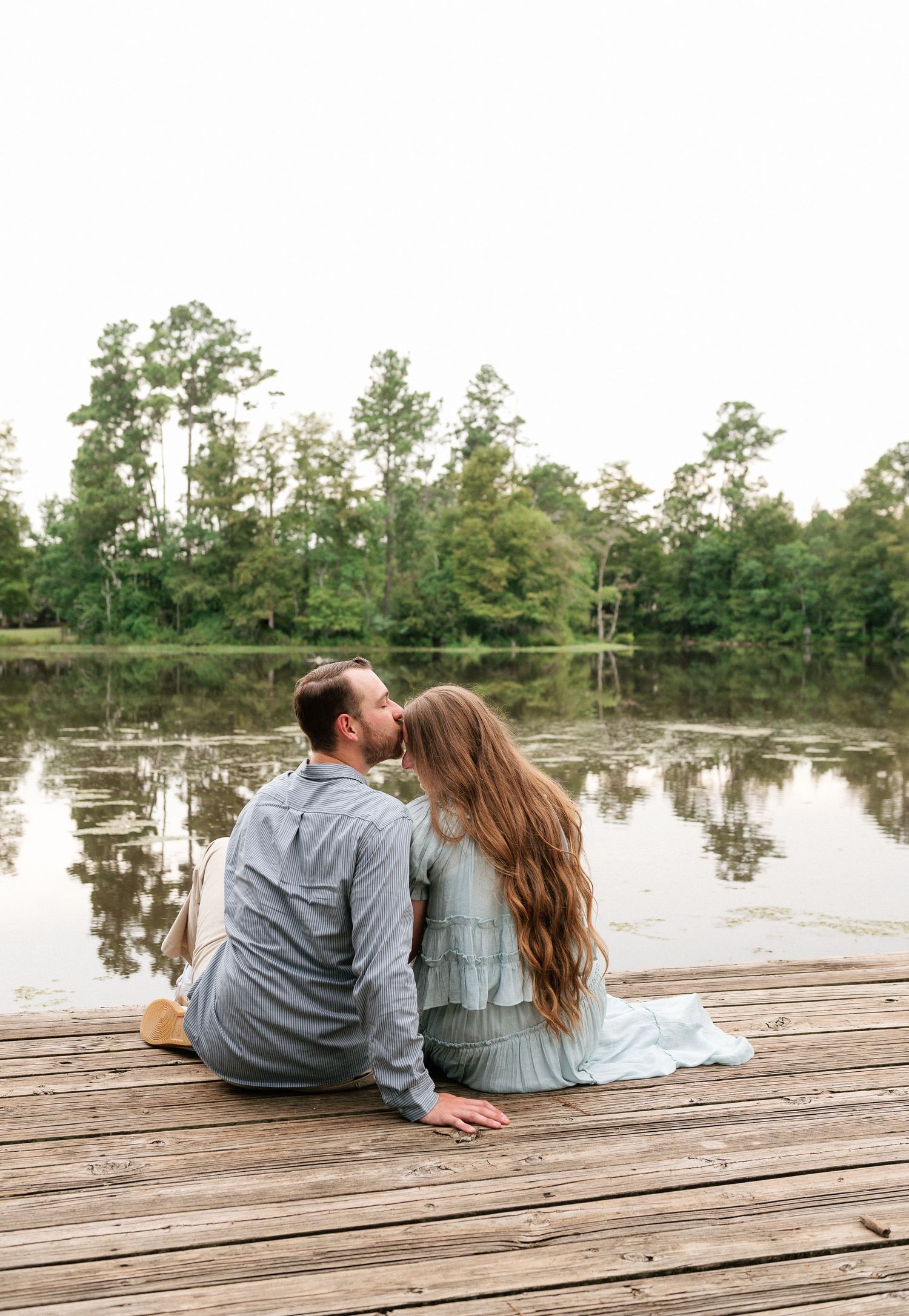 Couple sitting on a wooden dock by a calm lake, sharing a kiss surrounded by lush green trees.