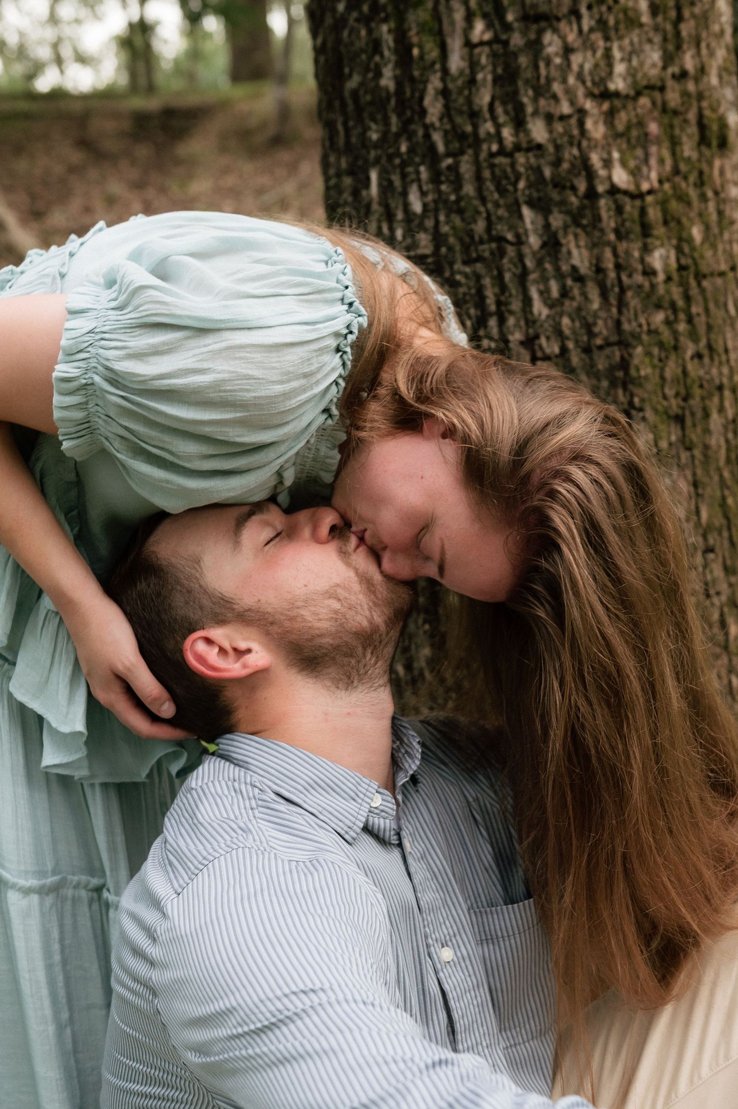 A couple shares a tender upside-down kiss leaning against a tree, woman in mint dress with long hair.