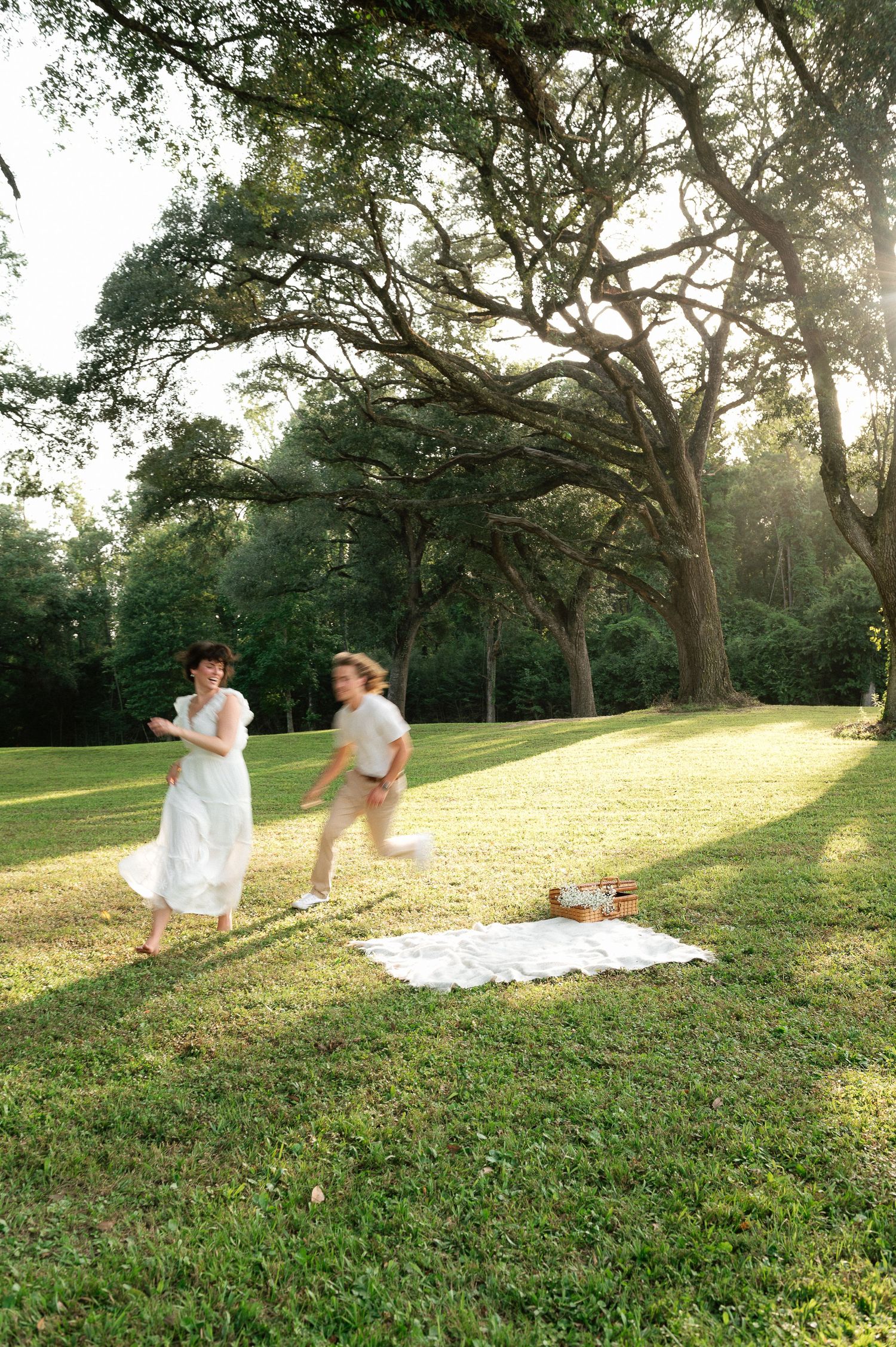 Two children in white outfits run playfully on lush green grass near a picnic blanket under large oak trees.