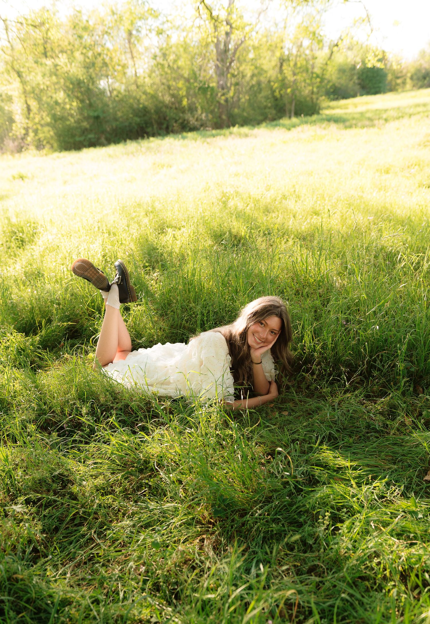 Young woman in white dress lying in lush green field, smiling, with legs raised, wearing dark shoes on sunny day.