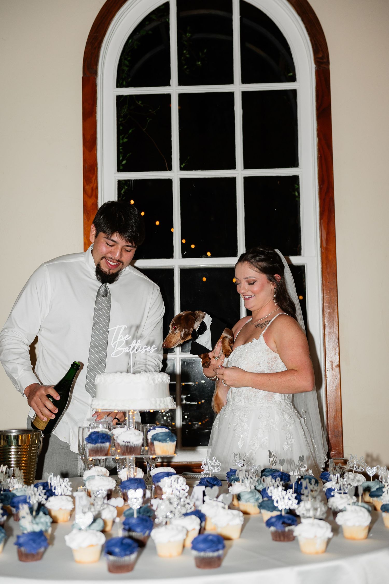 Bride and groom cutting wedding cake surrounded by navy blue cupcakes at elegant indoor reception.