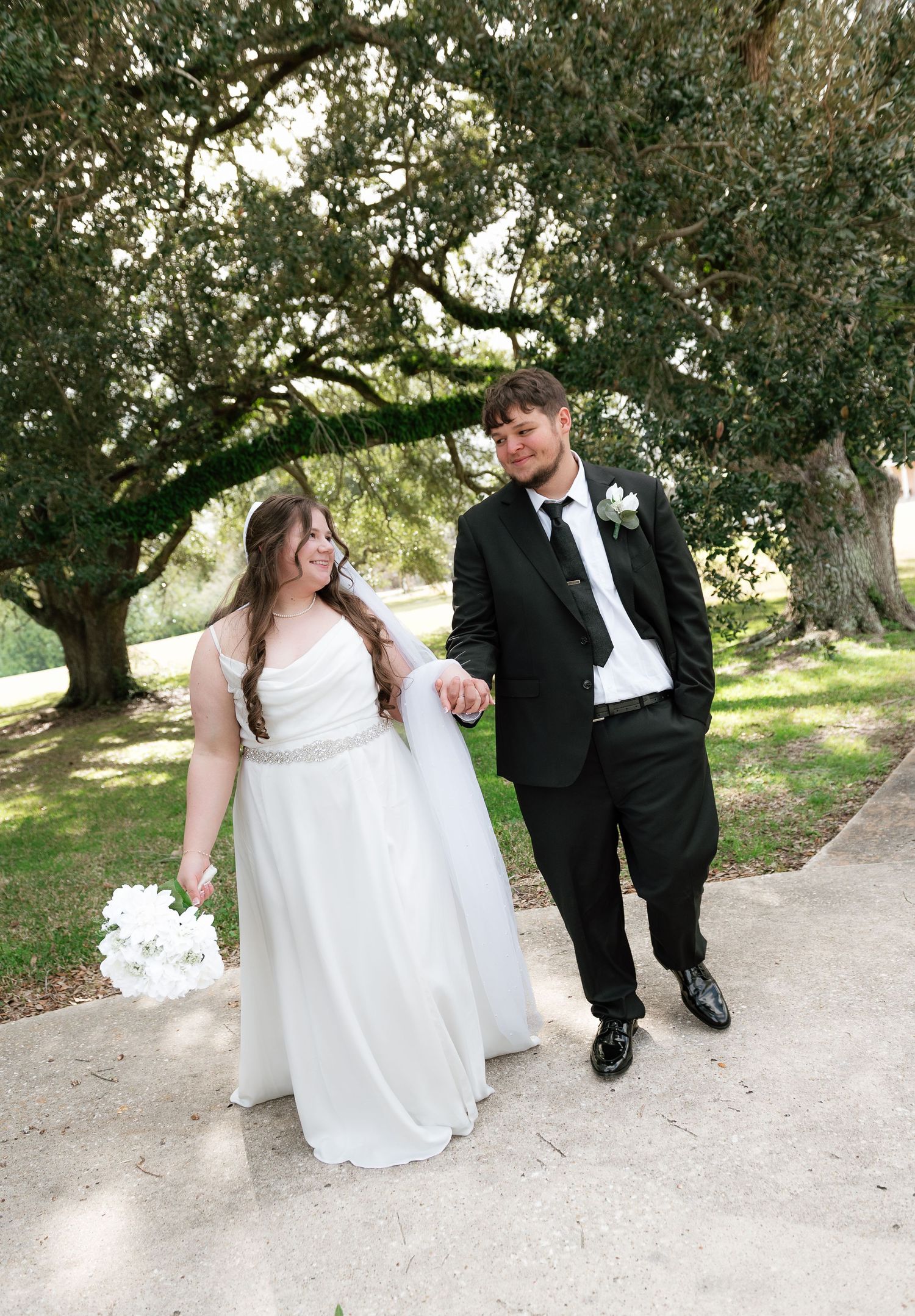 Bride in white gown and groom in black tux walk hand-in-hand on a garden path under lush oak trees.