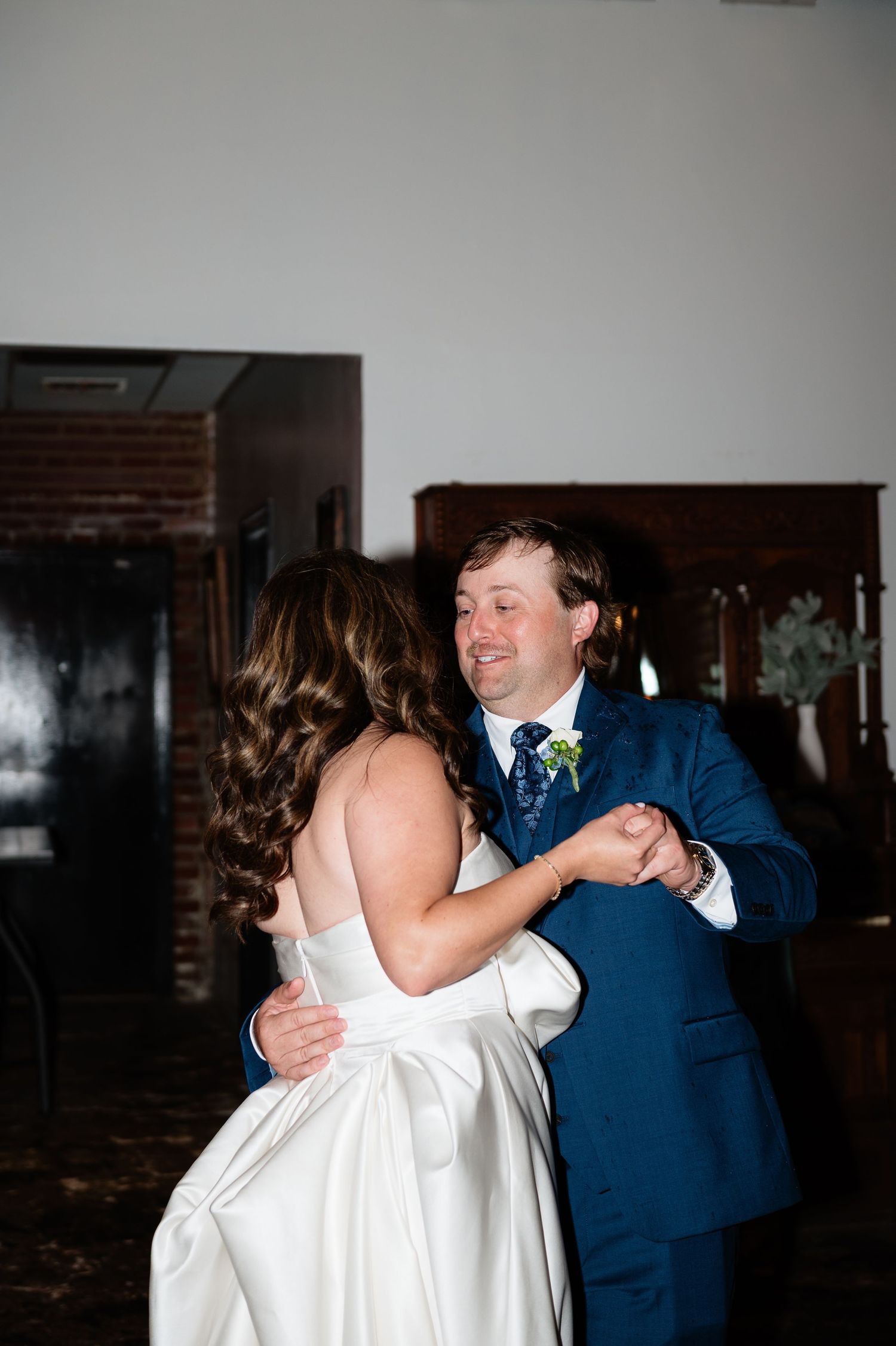 Bride and groom share their first dance, with groom in navy suit and boutonniere smiling warmly.