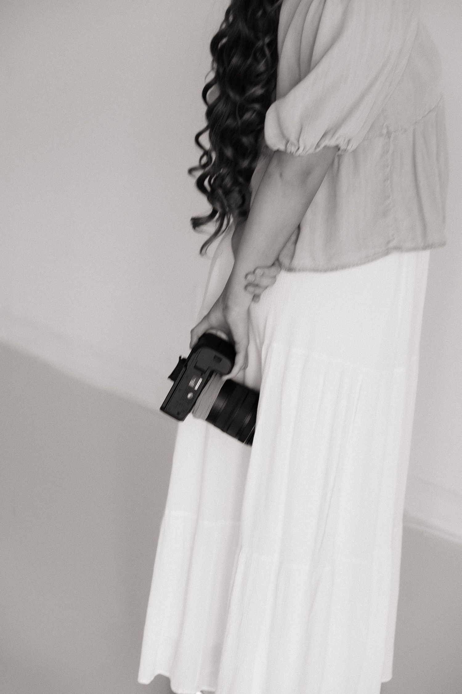 Black and white photo of a woman in a white skirt holding a camera behind her back, with long curly dark hair.