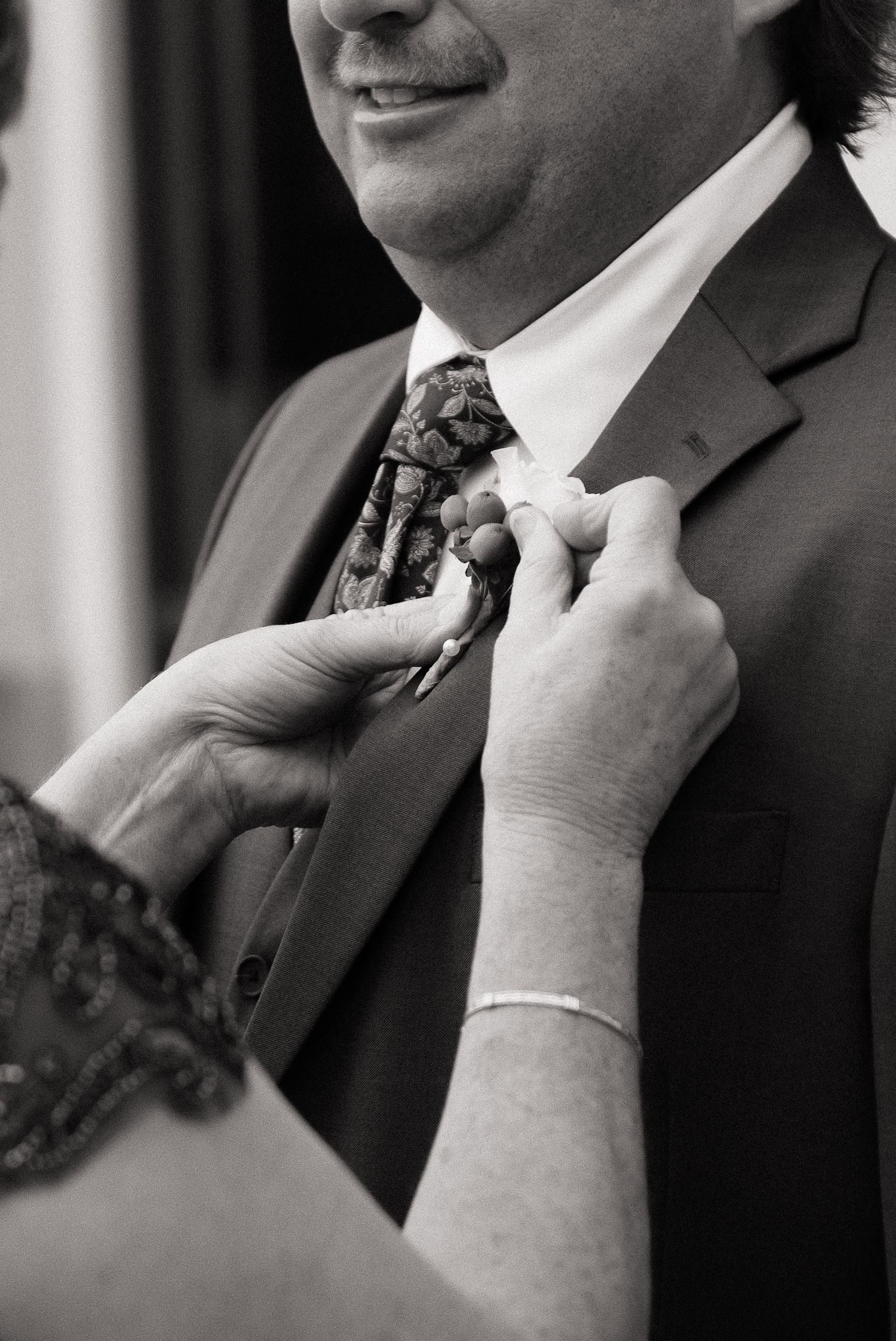 Black and white photo of hands adjusting a patterned tie on a suited man at a formal event.