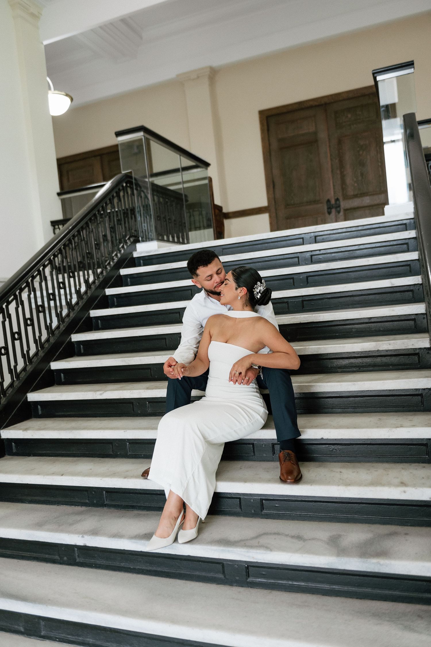 Bride and groom sitting together on elegant marble staircase, sharing a romantic moment in a classic indoor venue.