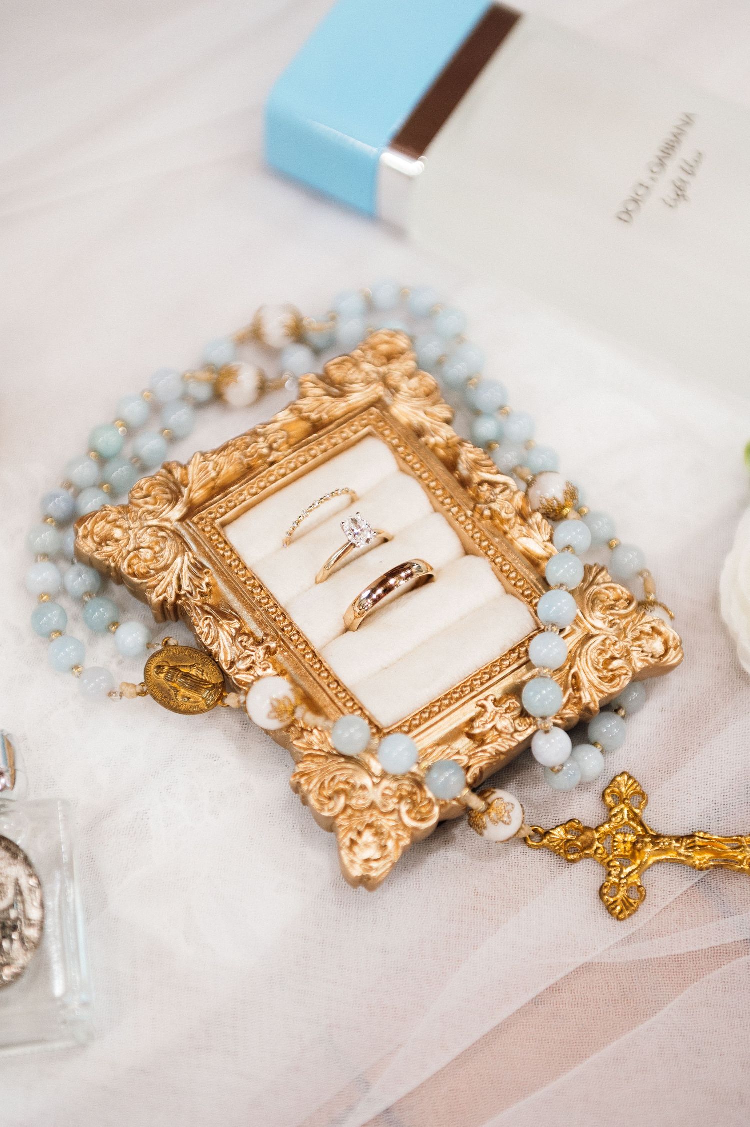 Gold ornate frame with wedding rings, blue rosary beads, and cross on white fabric near Dolce Gabbana perfume.
