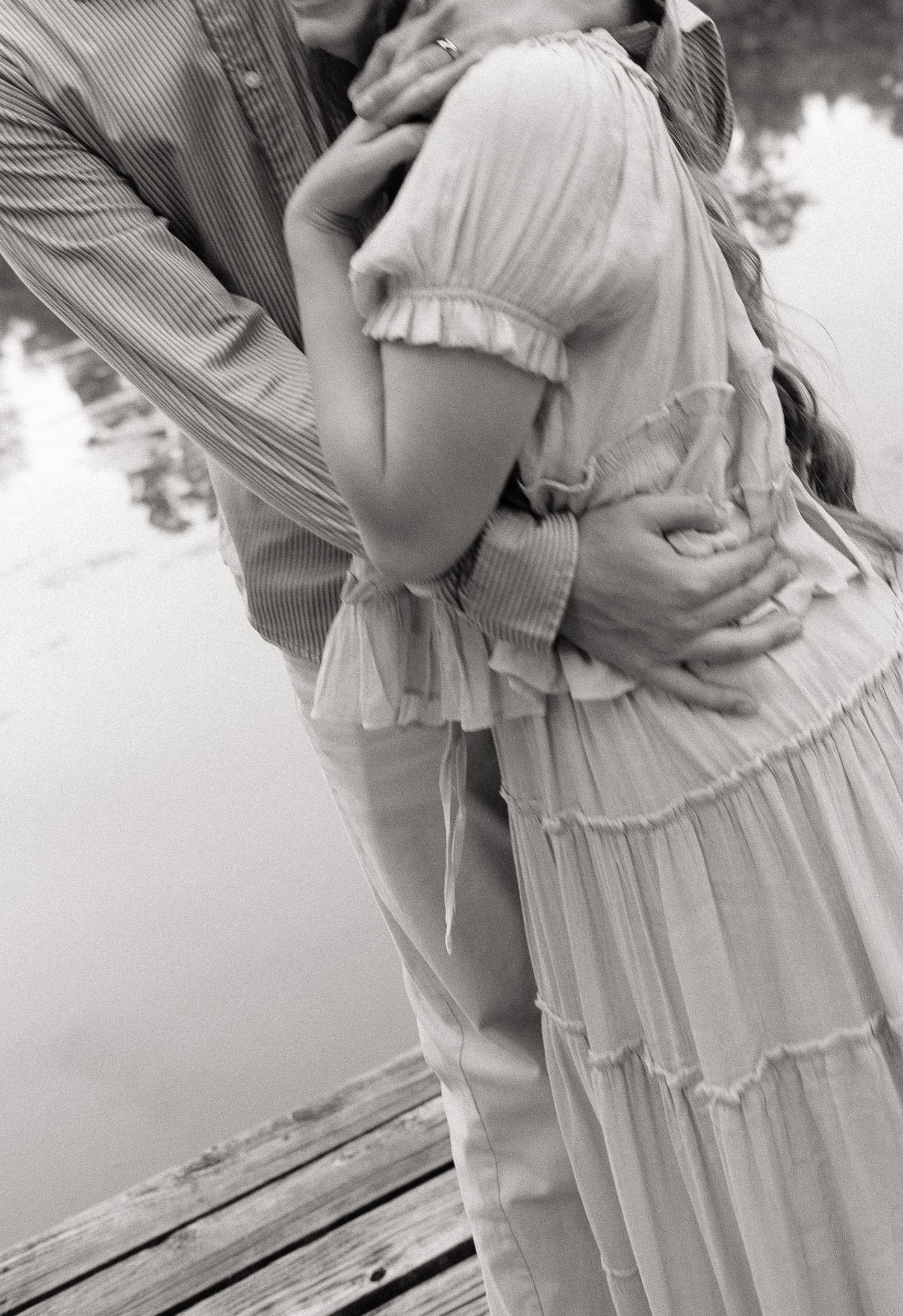 Black and white photo of a couple embracing on a wooden dock near a lake, woman in ruffled dress.