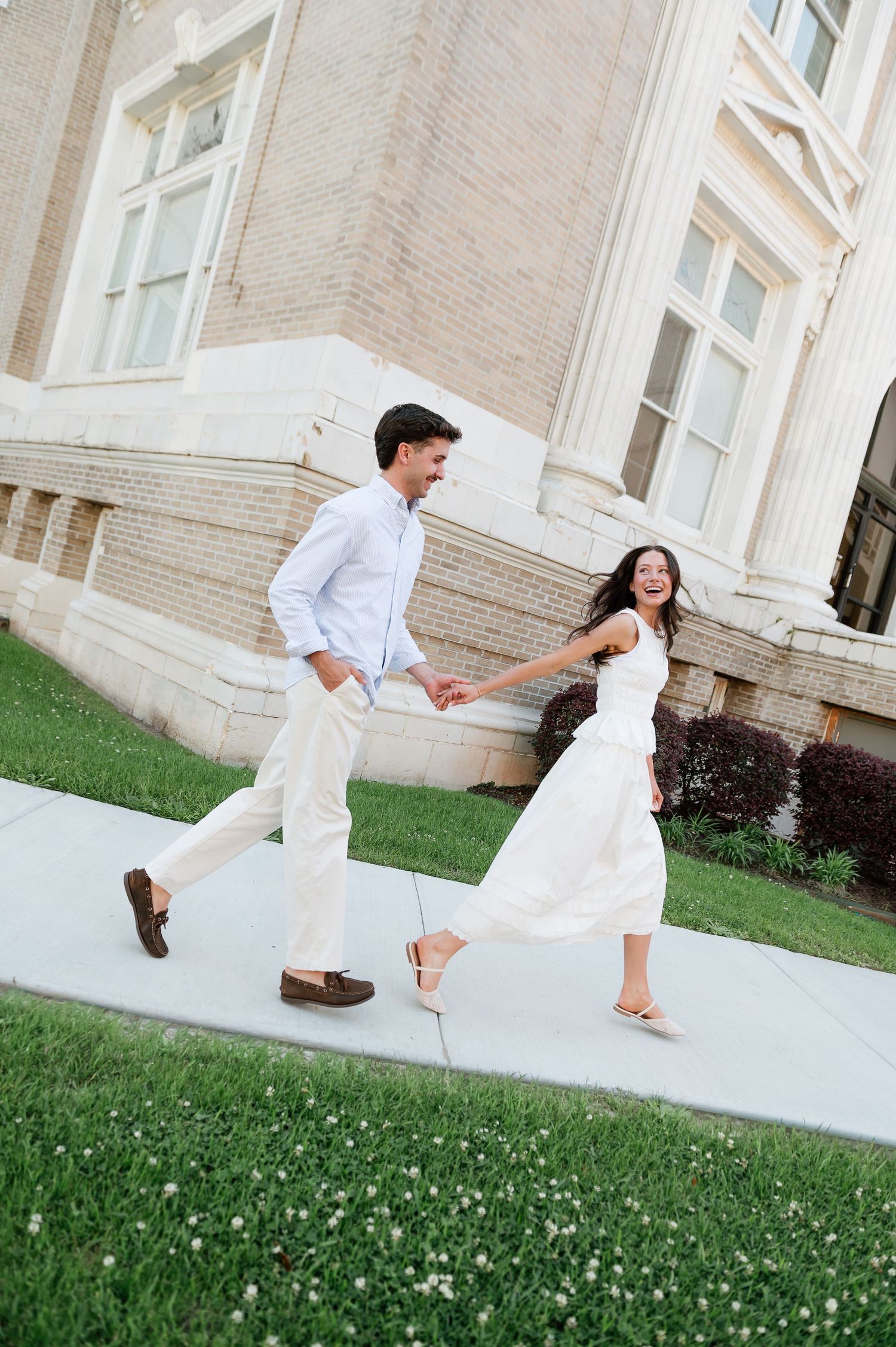 Happy couple in white outfits holding hands and walking along a sidewalk outside an elegant building.