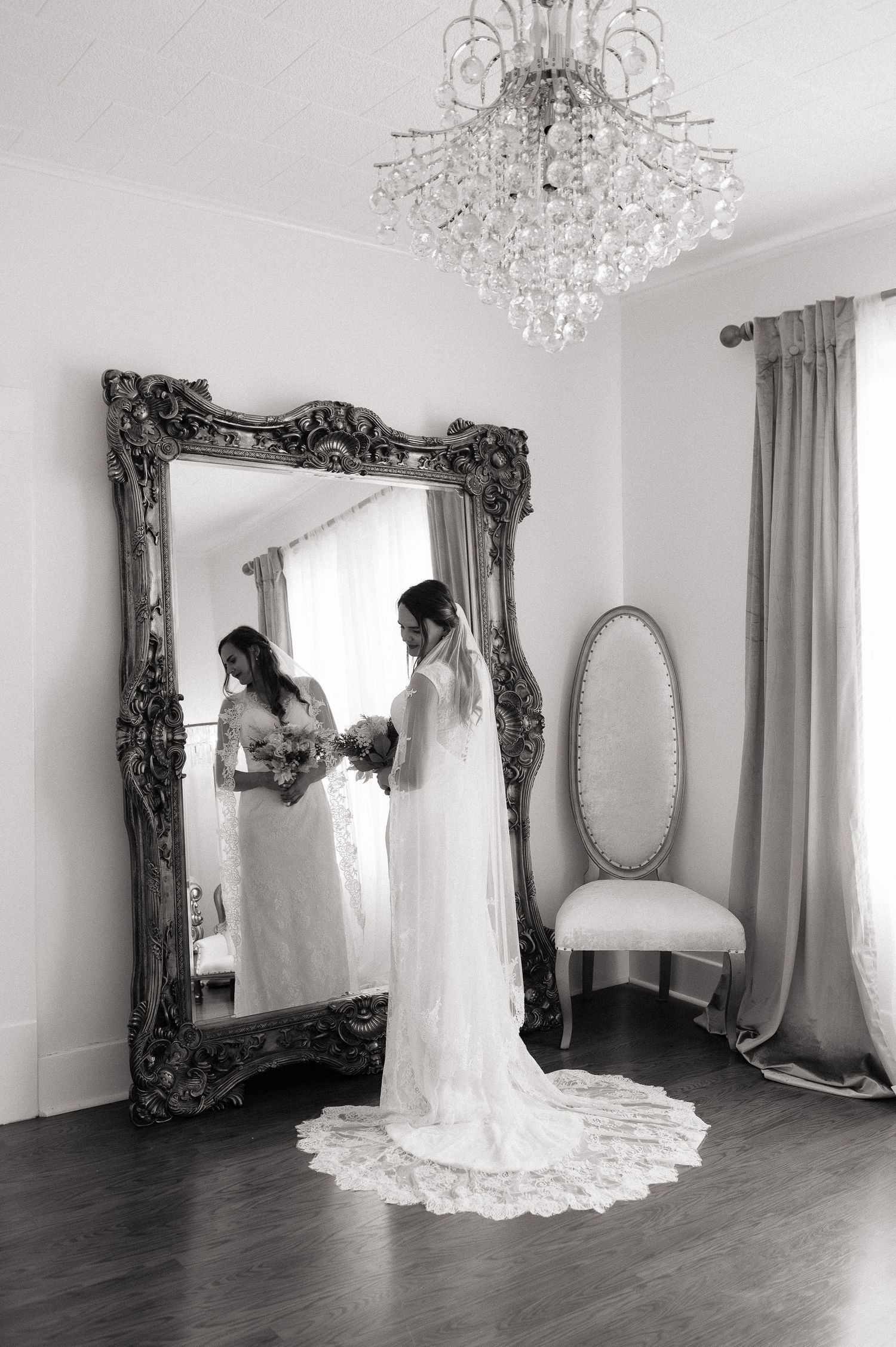 Bride in white gown and veil holding bouquet, reflected in ornate floor mirror beneath crystal chandelier.