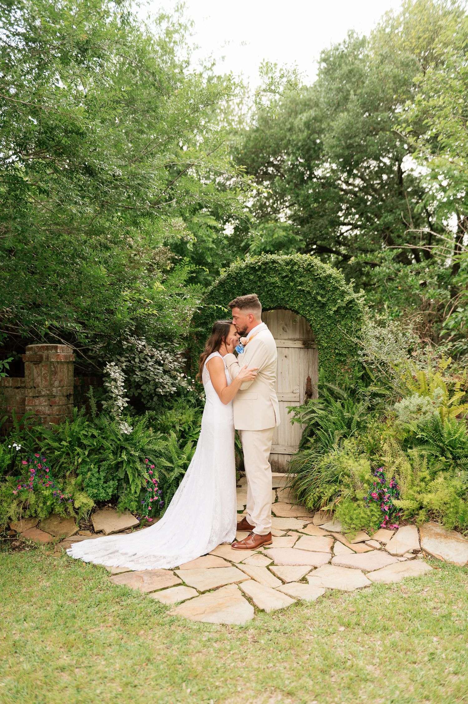 Bride and groom share a romantic embrace in a lush garden with a vine-covered stone archway on their wedding day.