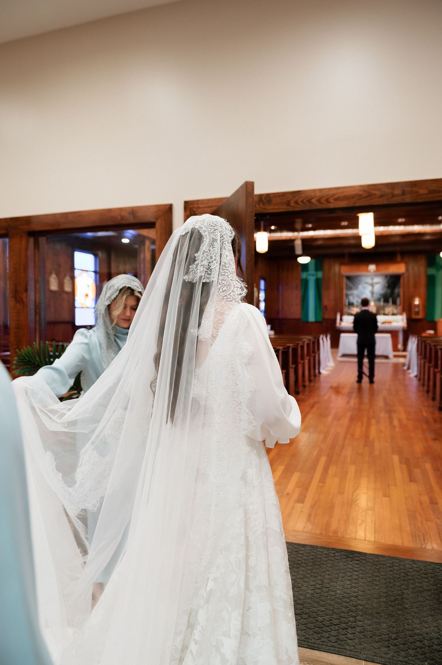 Bride in white lace gown and cathedral veil stands at church entrance, assisted by attendant before walking down aisle.