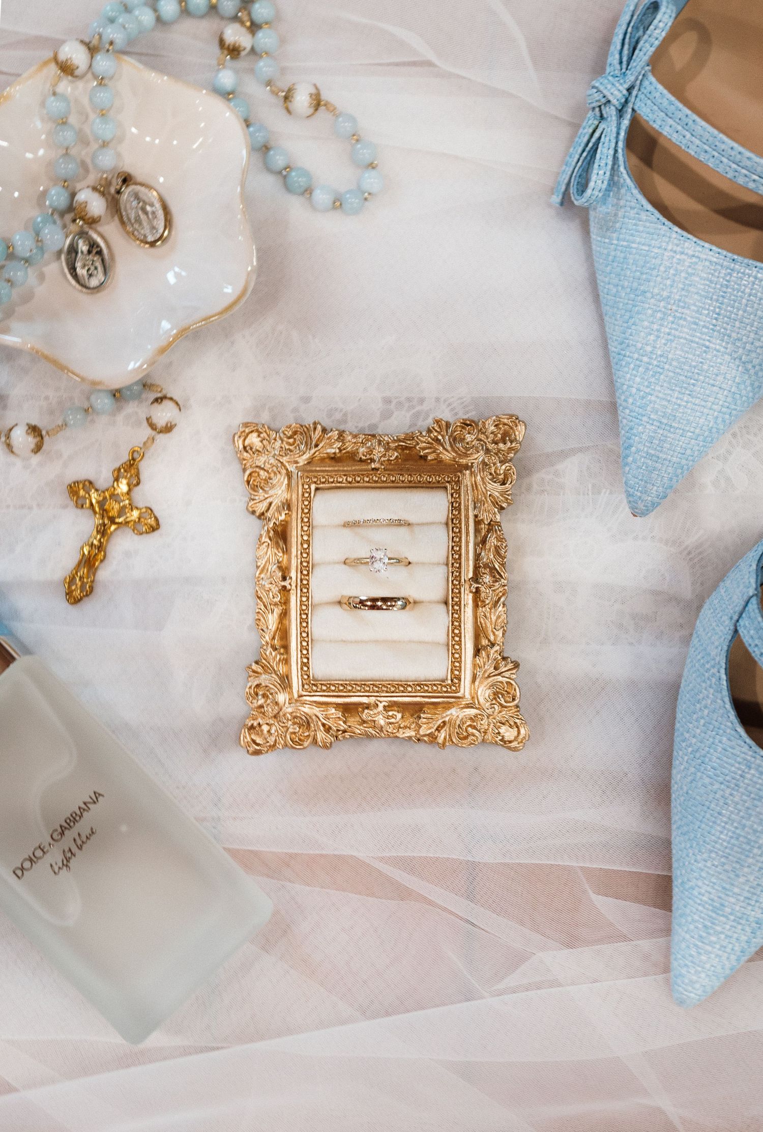 Gold ornate frame displaying wedding rings beside blue heels, rosary beads, crucifix, and Dolce Gabbana perfume.