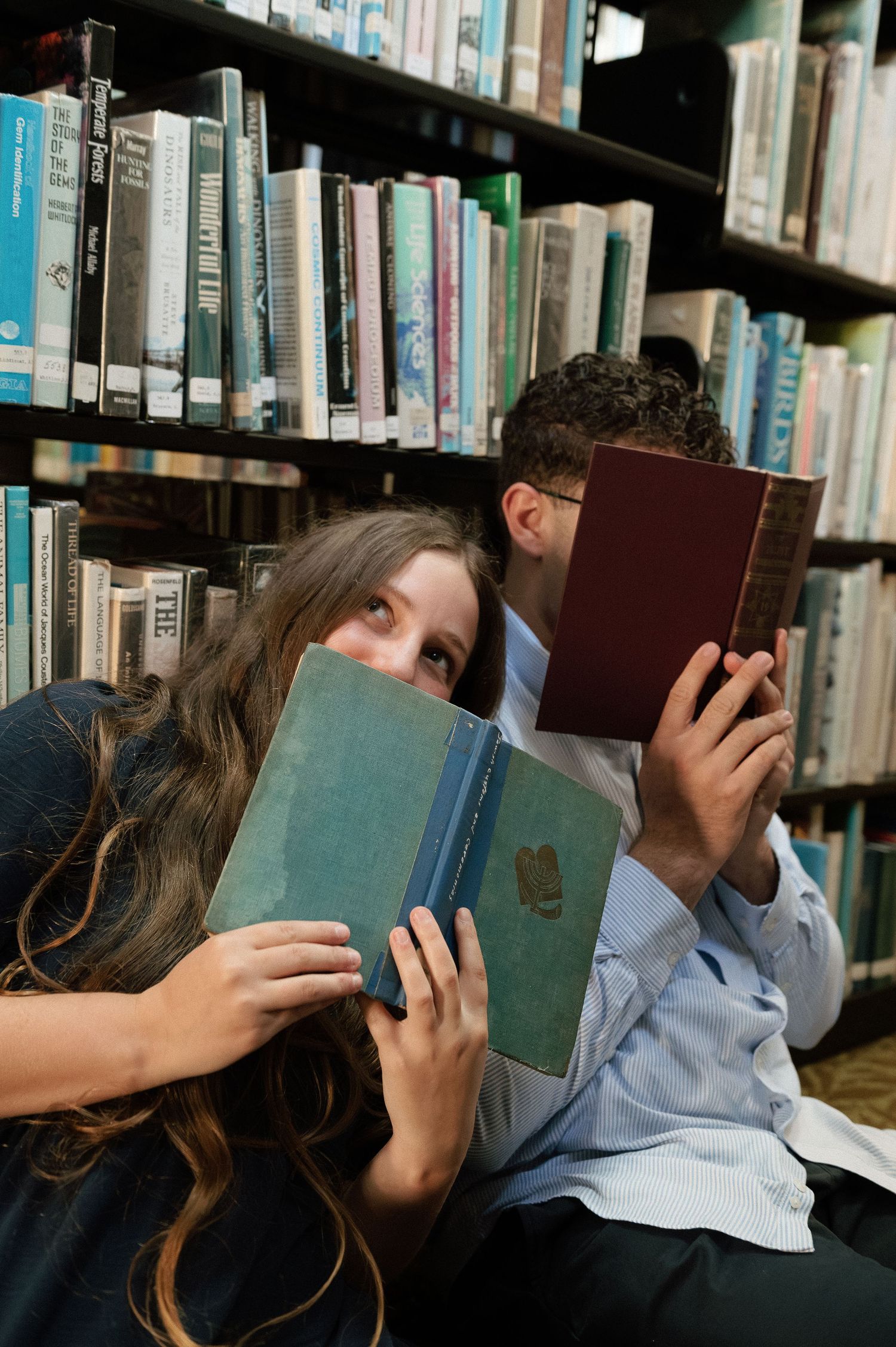 Two students sit against library shelves, playfully hiding their faces behind vintage hardcover books.