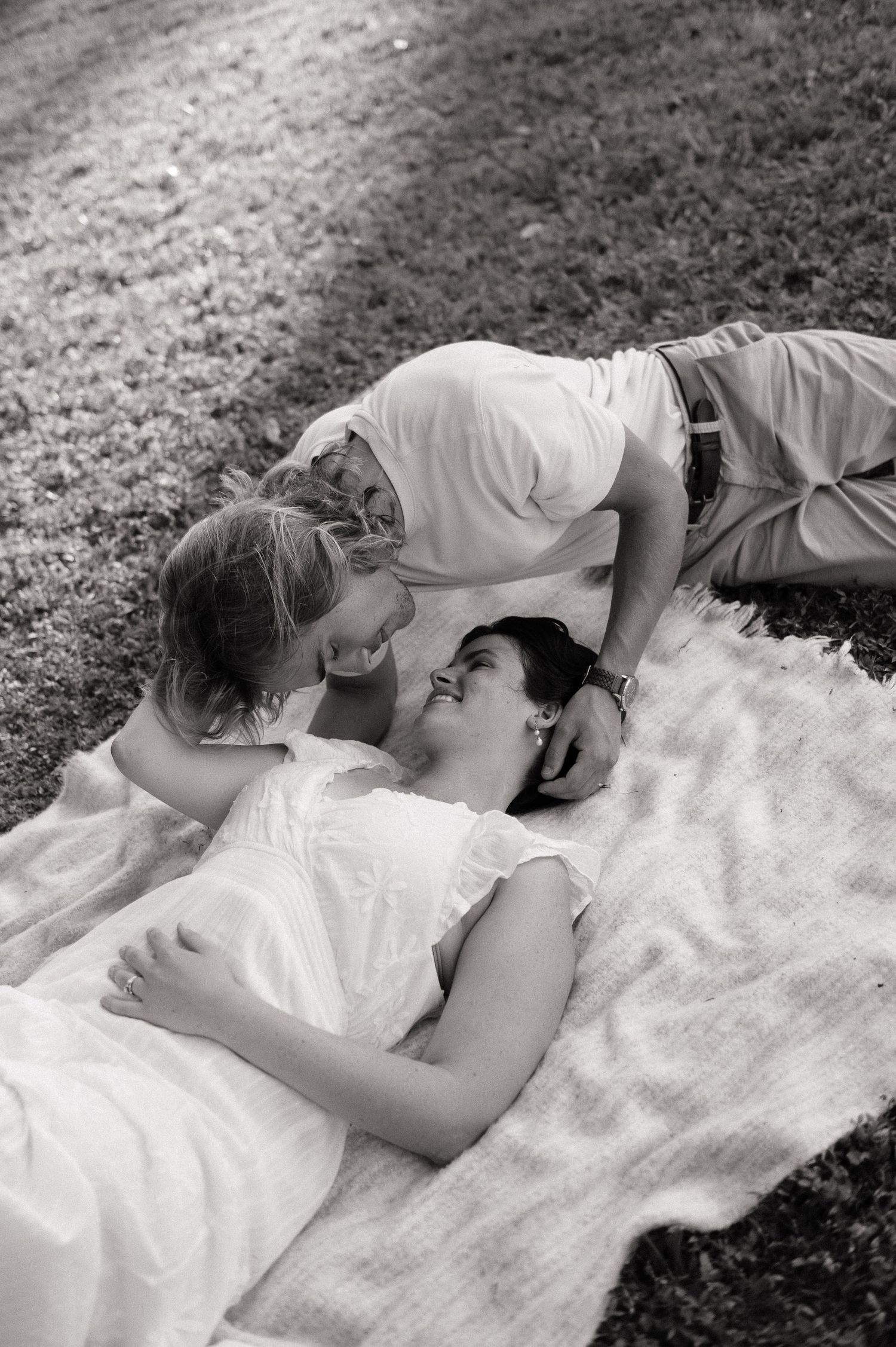 Black and white photo of a couple lying on a blanket in grass, sharing a tender romantic moment outdoors.
