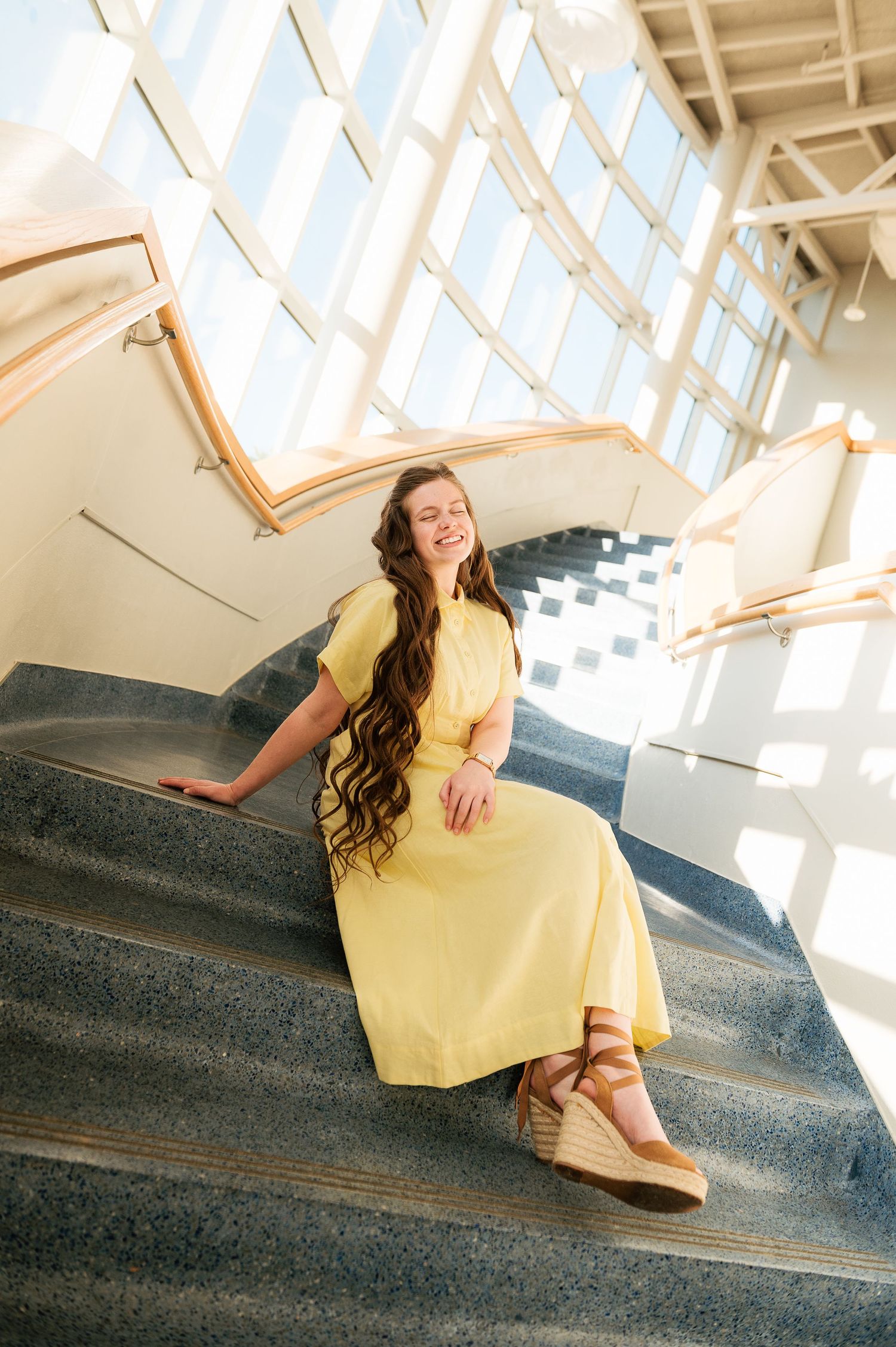 Young woman in yellow maxi dress with wavy hair sits smiling on sunlit interior staircase with large windows.