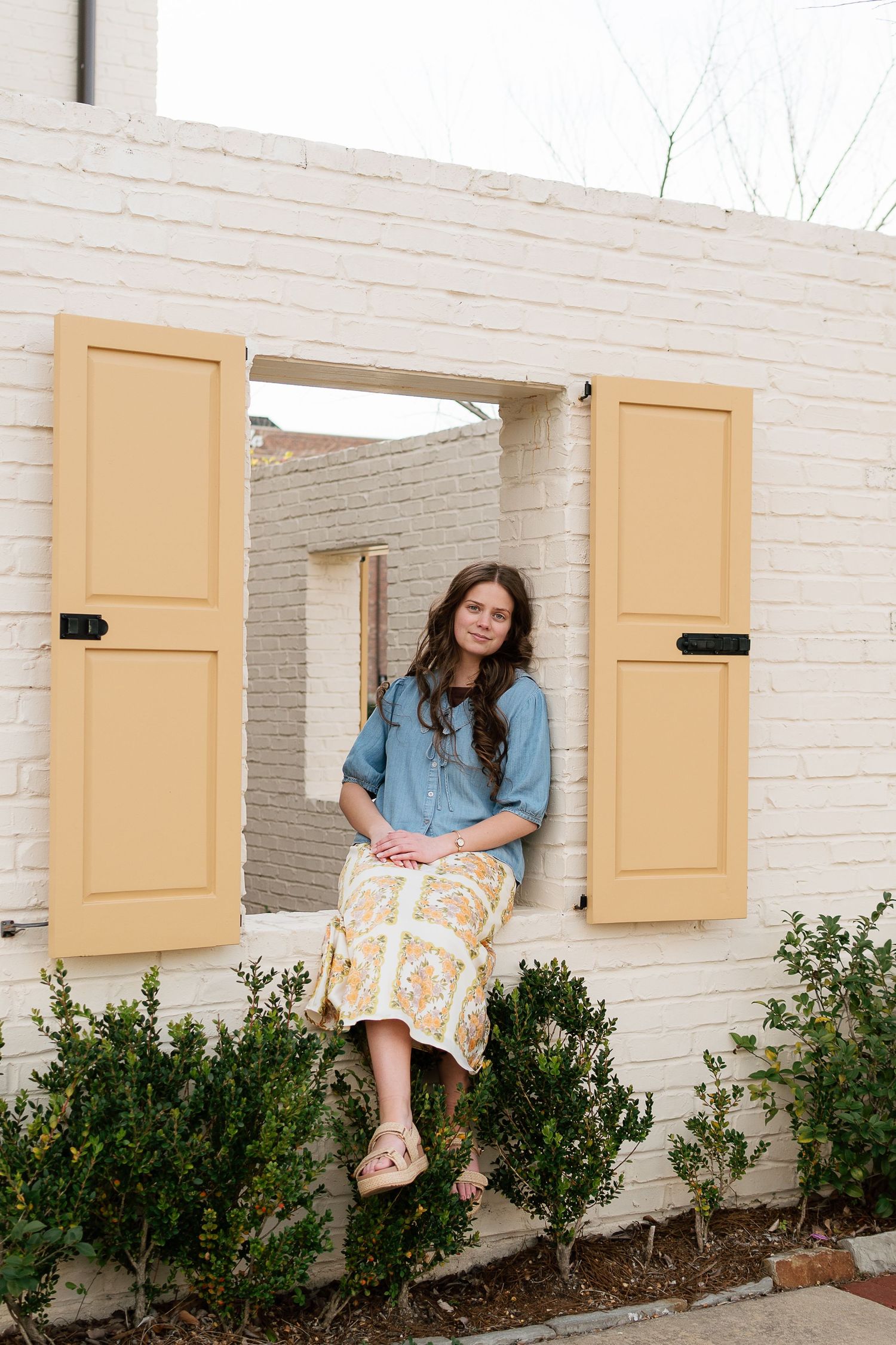 Woman in blue shirt and floral skirt sitting on white brick window ledge with yellow shutters.