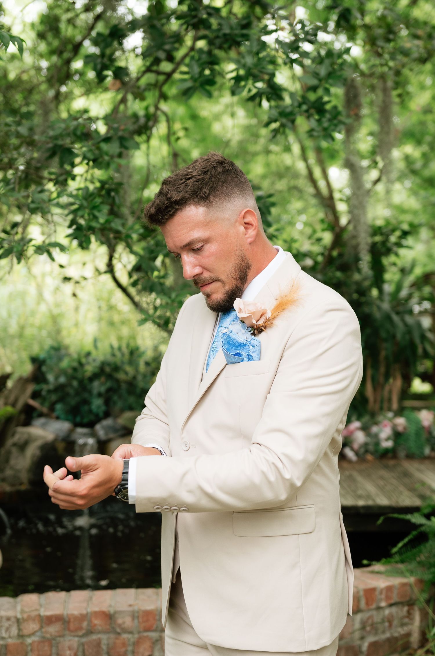 Groom in beige suit adjusting cufflinks outdoors, wearing blue pocket square and floral boutonniere in lush garden setting.