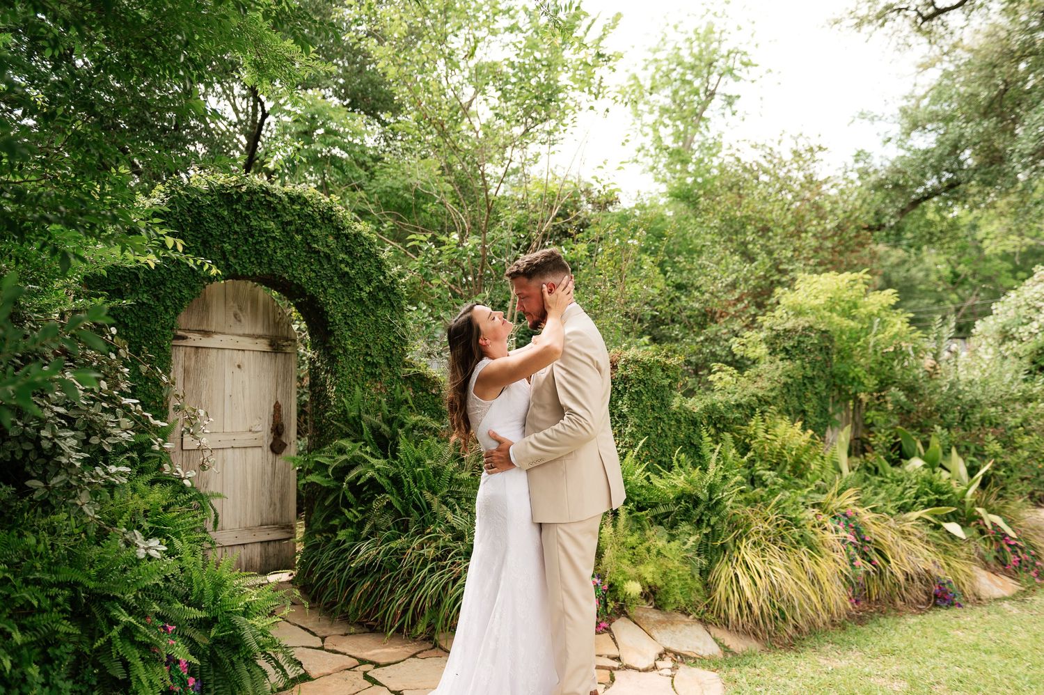 Newlywed couple sharing a romantic kiss in a lush garden with a rustic wooden door backdrop.