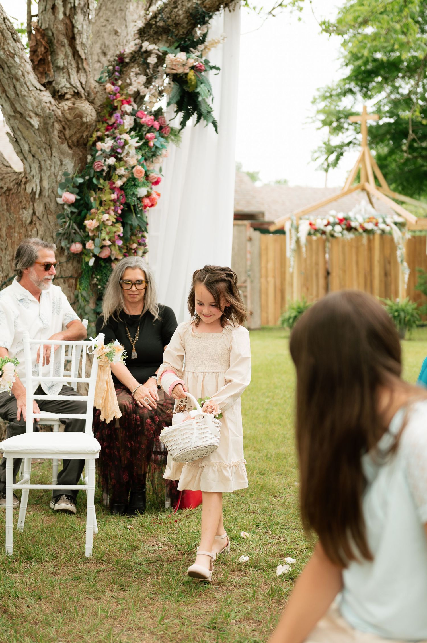A young flower girl in a white dress carries a basket down the aisle at an outdoor garden wedding ceremony.