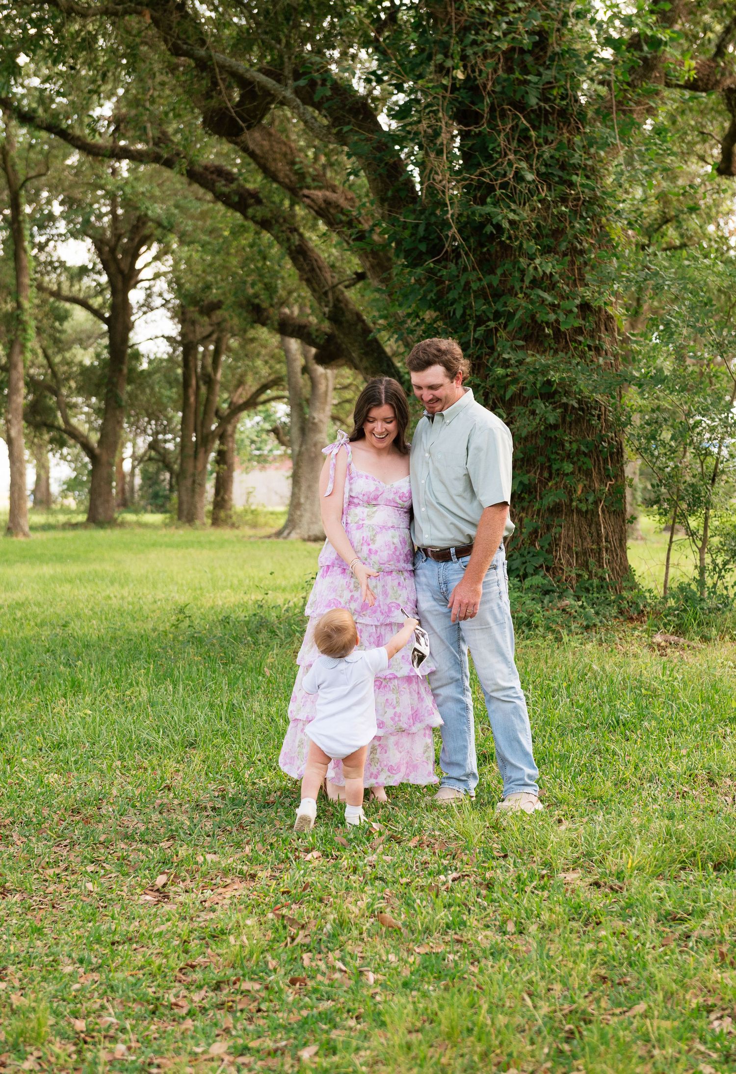 Young family with toddler enjoying outdoor maternity photo session among green trees on grassy lawn.