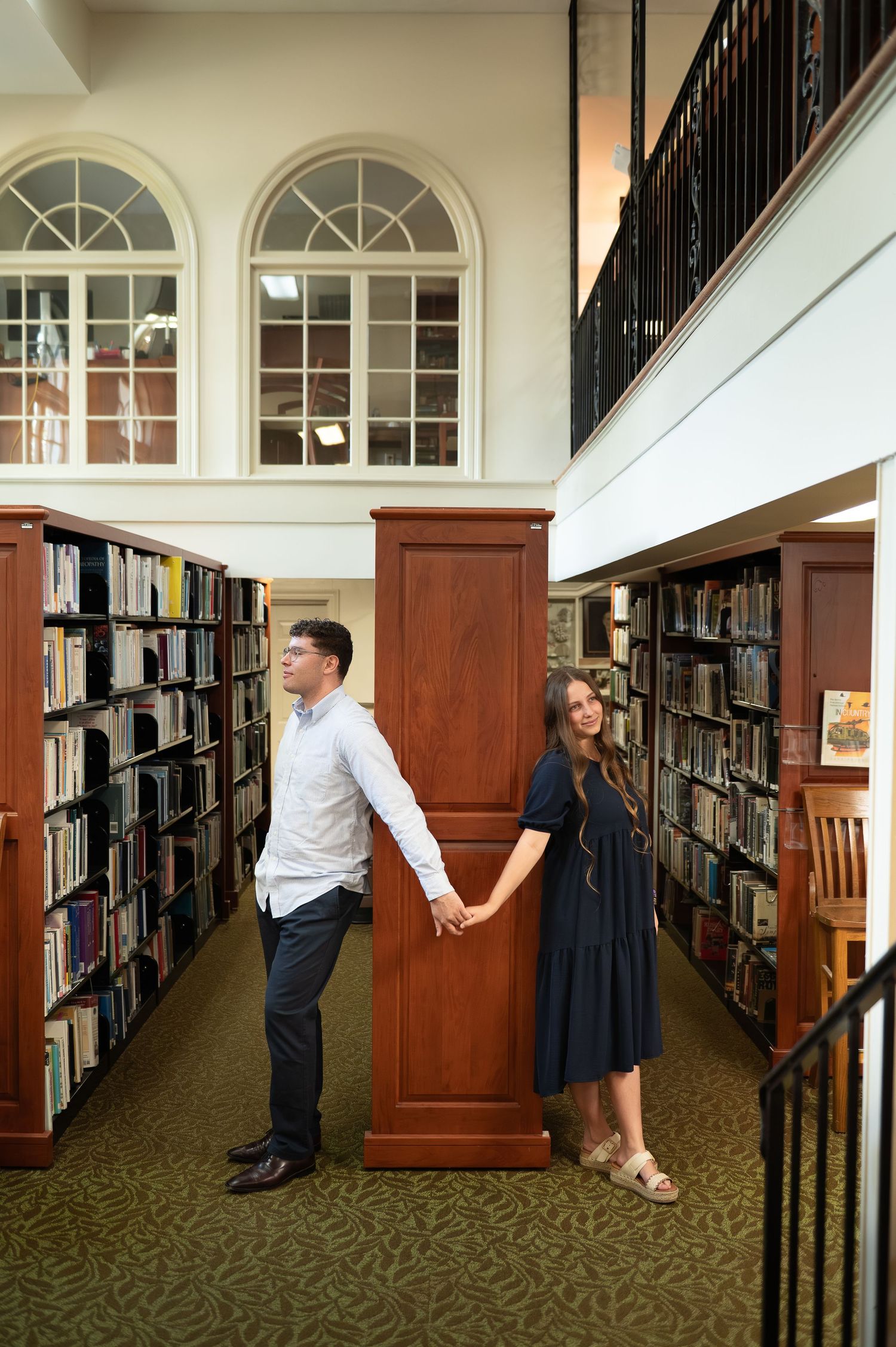 A couple holds hands between library bookshelves, man in white shirt and woman in navy dress, in a classic two-story library.