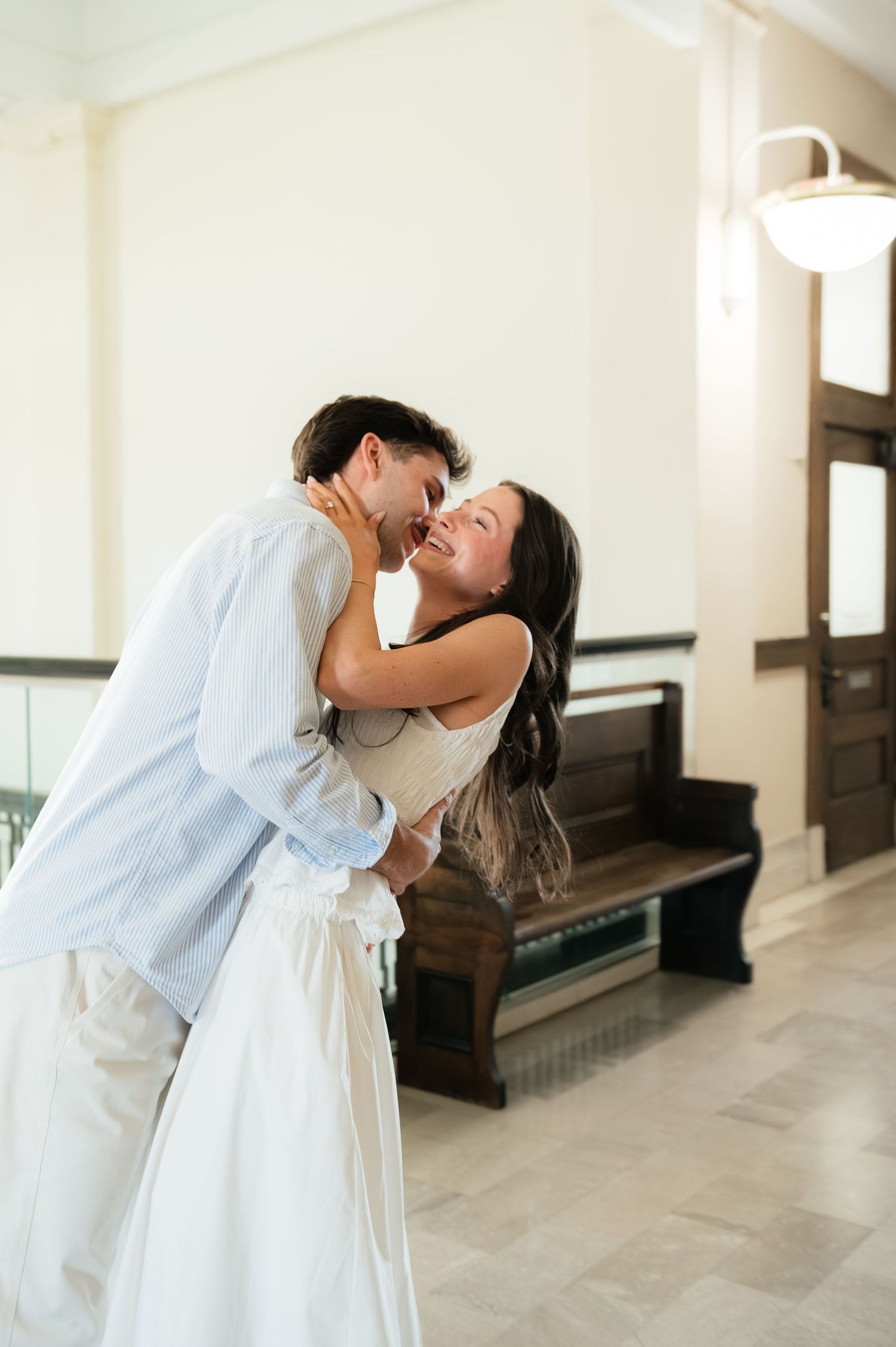 A joyful couple shares a romantic kiss in a bright indoor hallway, the woman in a white dress laughing with delight.