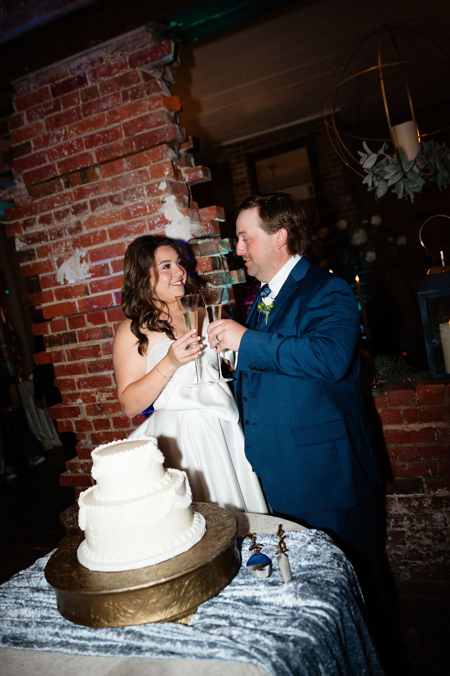 Bride and groom toasting champagne beside white three-tier wedding cake at reception.
