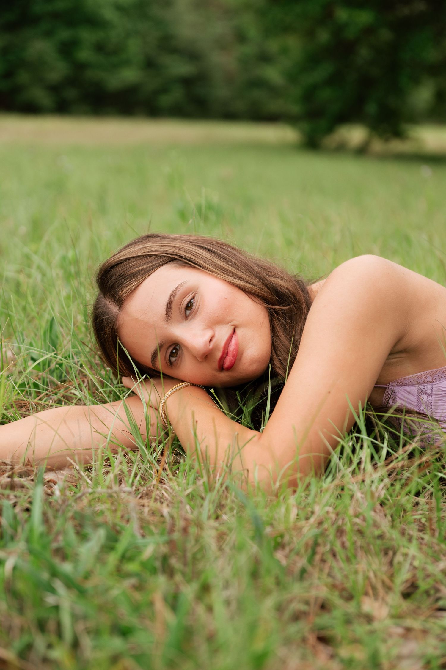 Young woman with long brown hair lying in green grass field, smiling at camera on a sunny day.