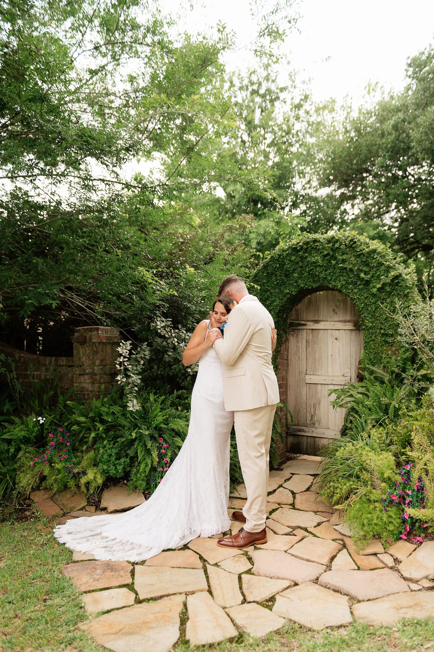 Bride and groom embrace near ivy-covered arched door on stone path in lush garden wedding setting.