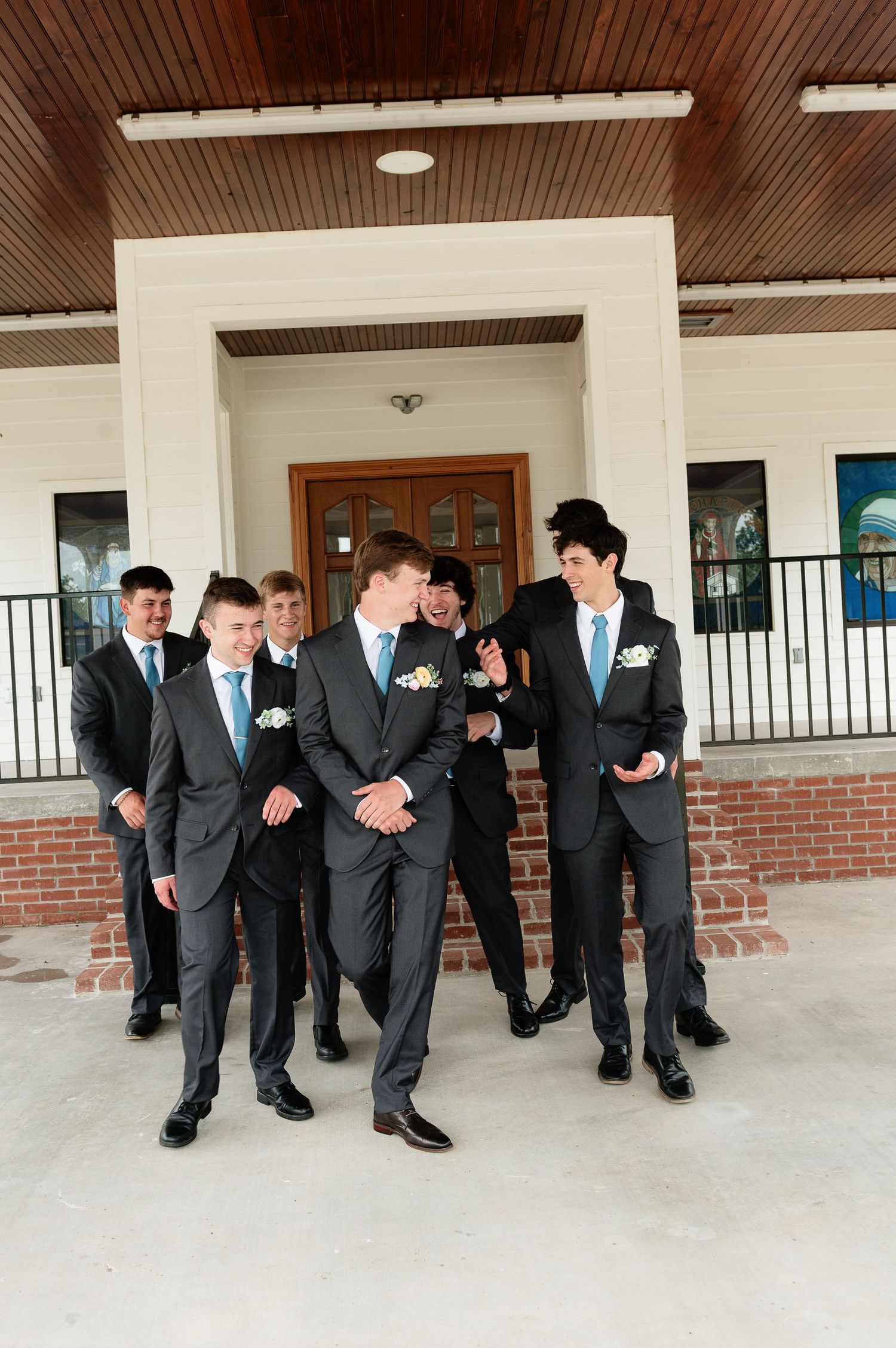 Group of young men in dark suits with light blue ties posing outside a brick building for a formal event.