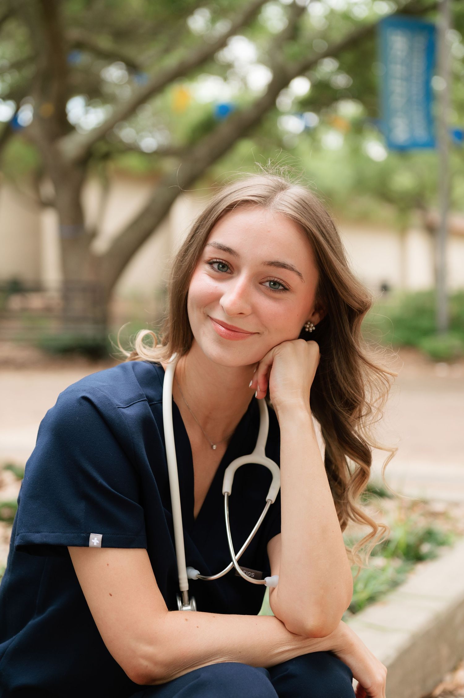 Young nursing student in navy scrubs with stethoscope smiles outdoors on a college campus.