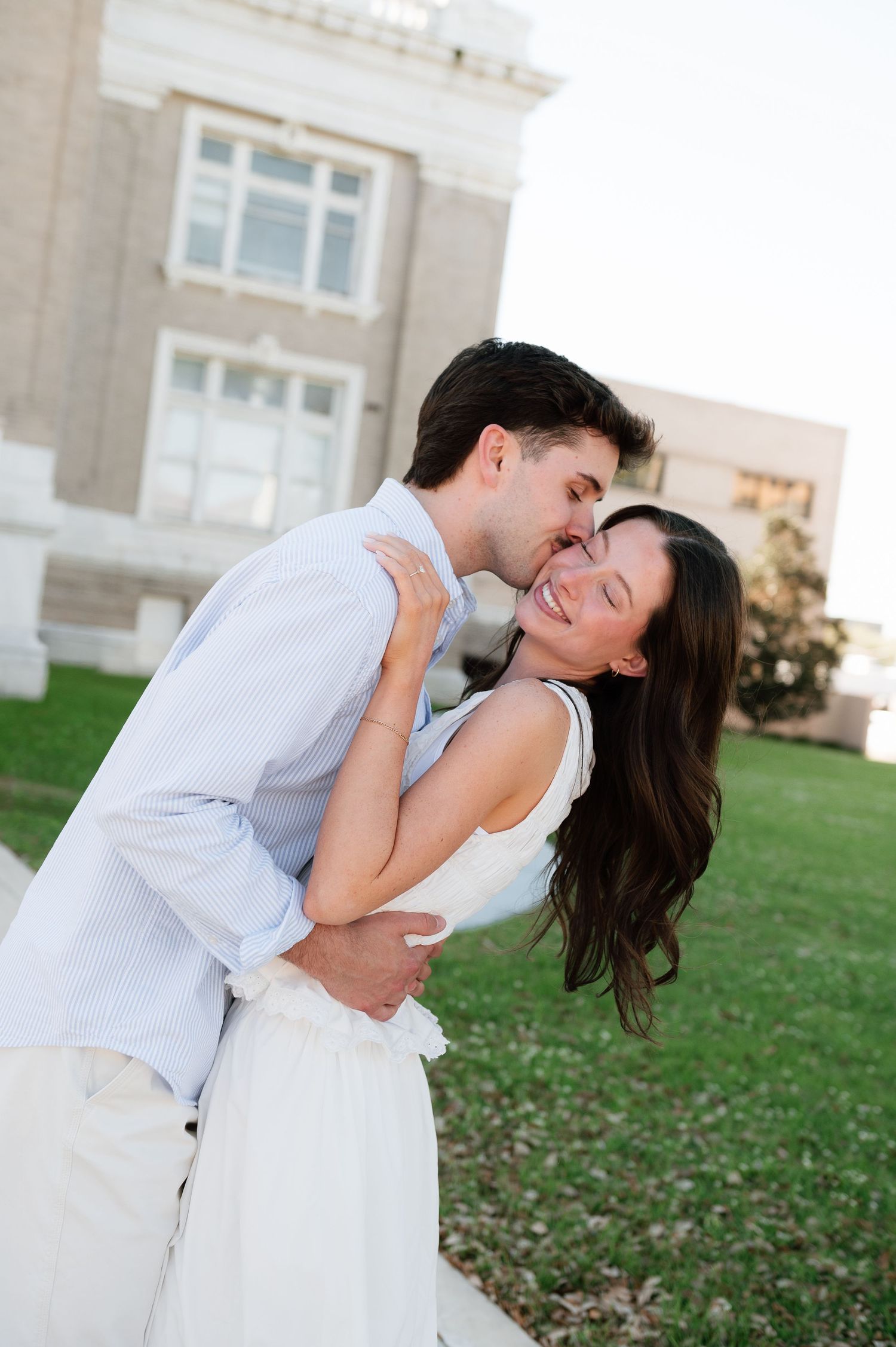 A couple shares a romantic kiss outdoors, with the woman in a white dress laughing joyfully near a stone building.