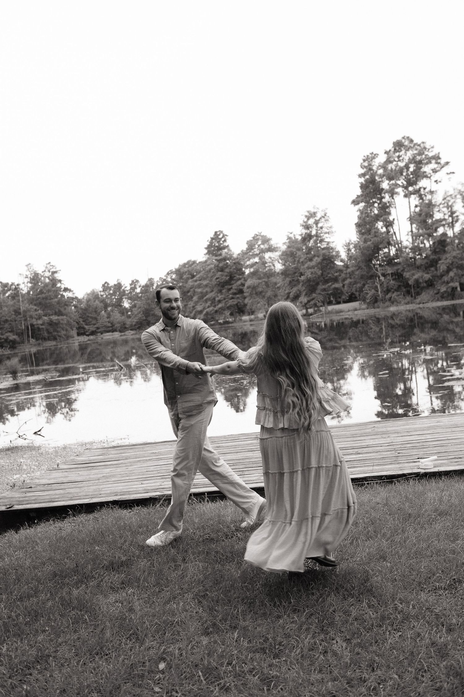 Black and white photo of a couple dancing on grass near a tranquil lakeside dock.
