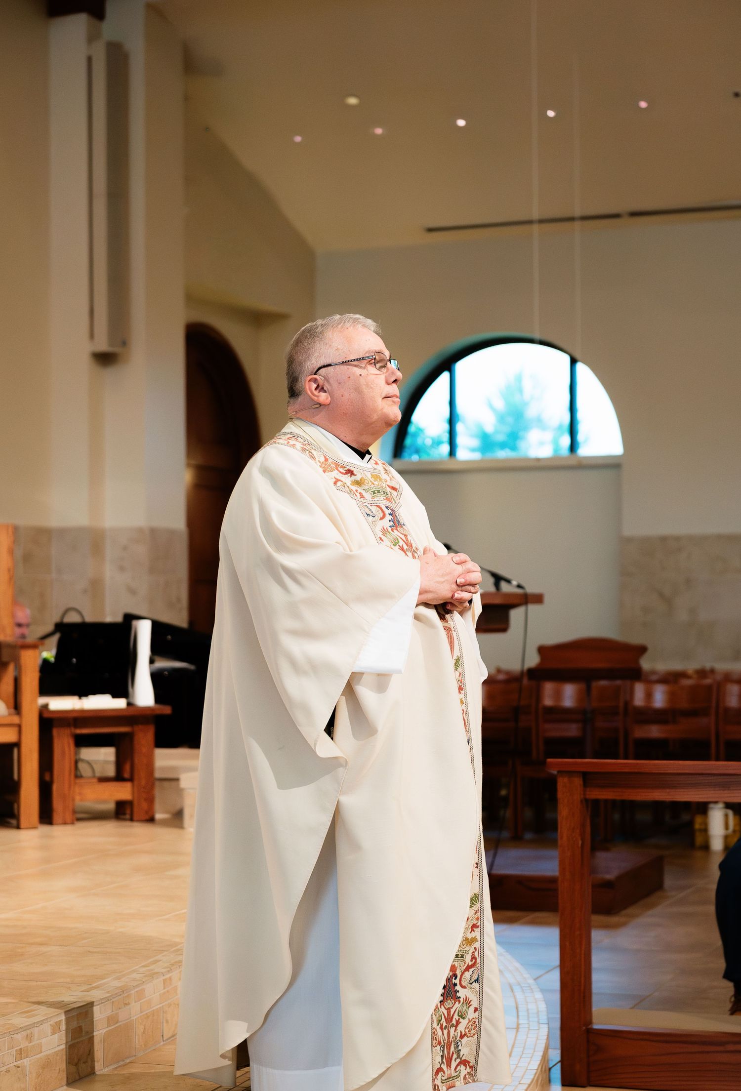Catholic priest in ornate white vestments standing at altar during wedding ceremony in modern church.