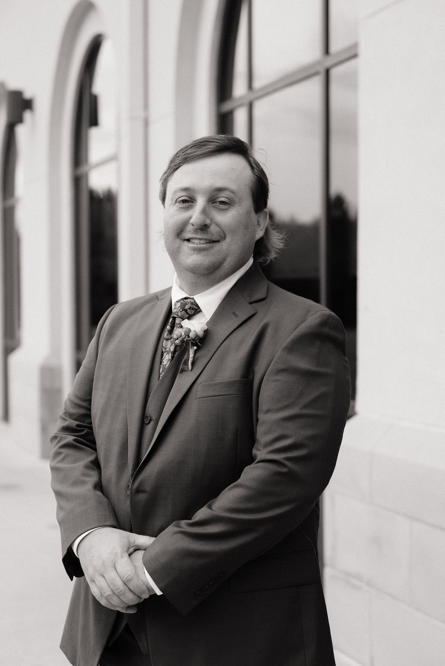 A smiling man in a suit and floral tie poses outdoors in a black and white professional portrait photo.