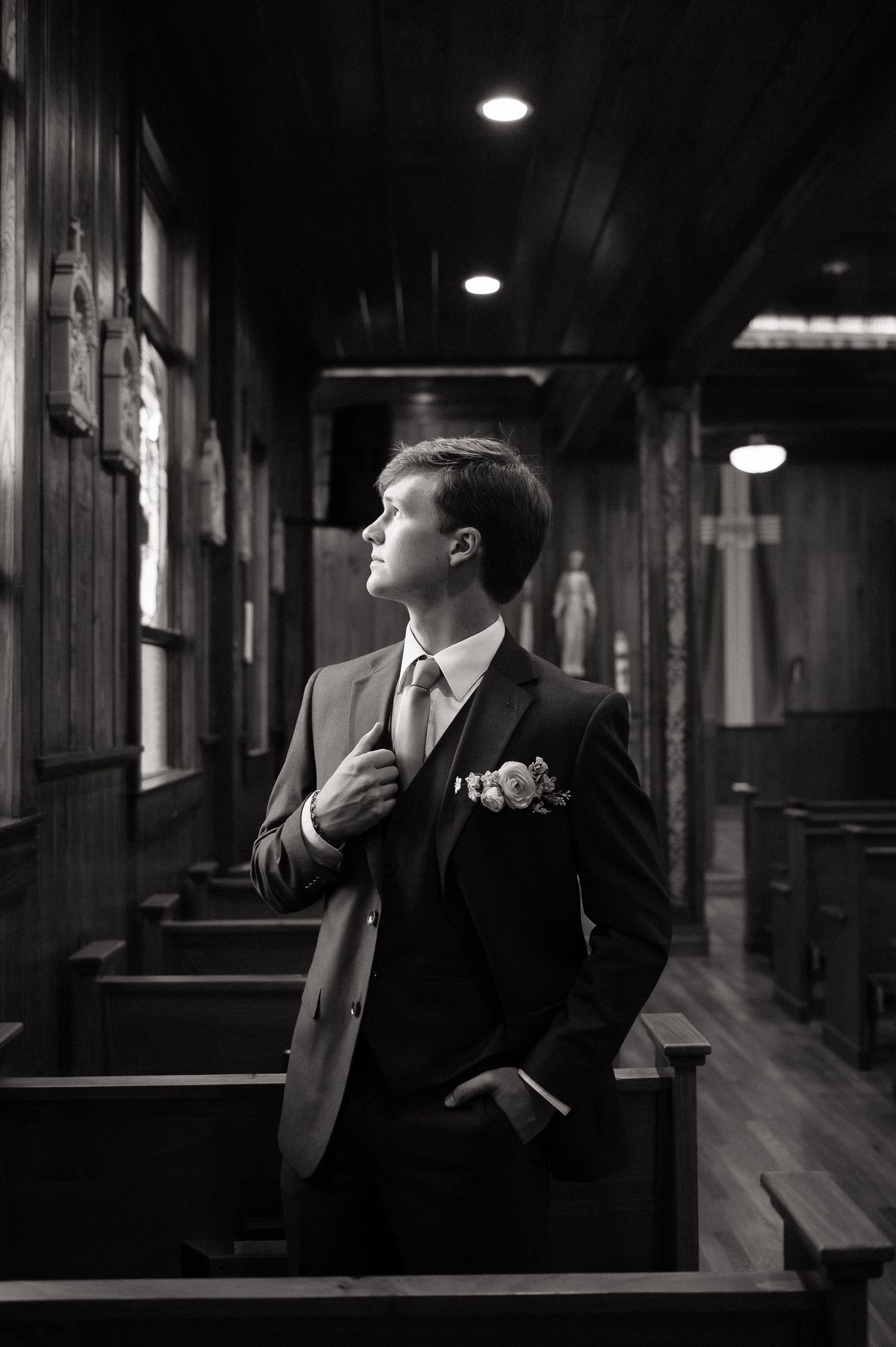 A groom in a dark suit with boutonniere adjusts his jacket while standing in a wooden church aisle, black and white photo.