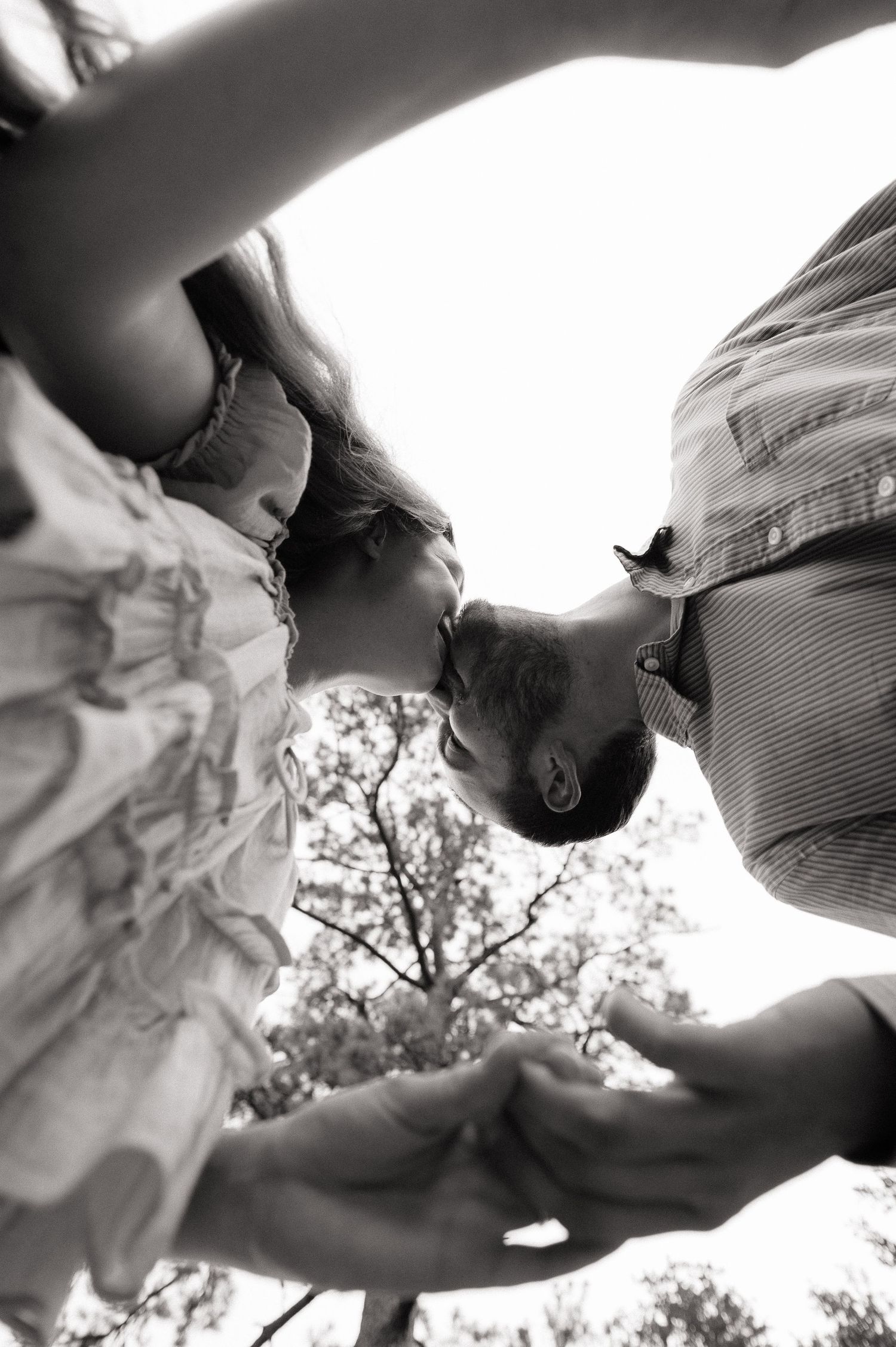 Black and white photo of a couple kissing, shot from below with trees in the background.