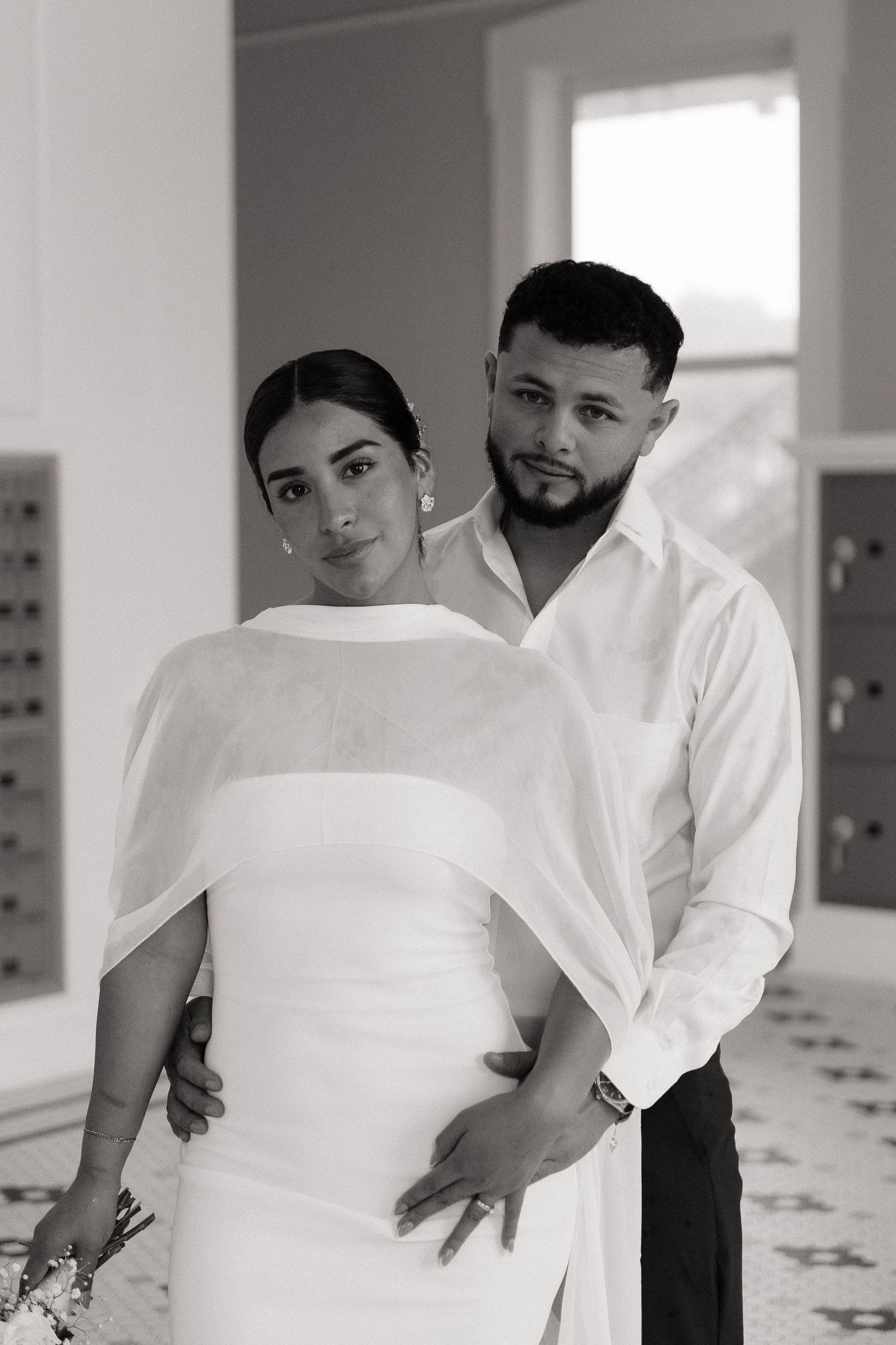 Black and white wedding photo of a couple posing elegantly, bride in white gown with cape, groom in white shirt.