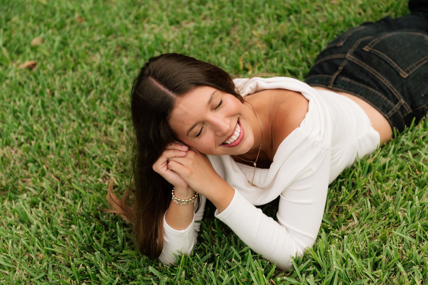 A young woman with long brown hair laughs joyfully while lying on lush green grass outdoors.