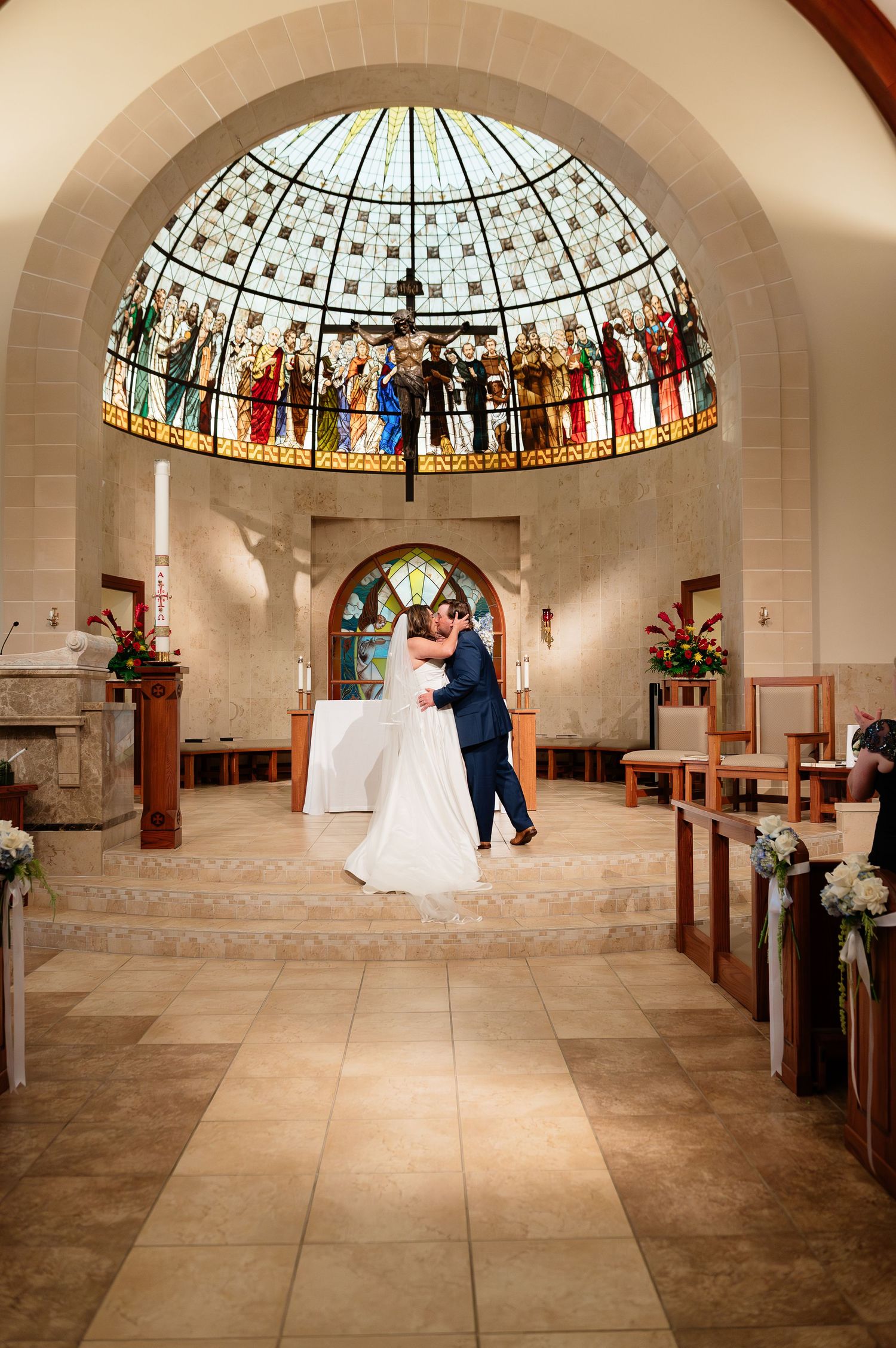 Bride and groom share first kiss under stunning stained glass dome in elegant church ceremony.