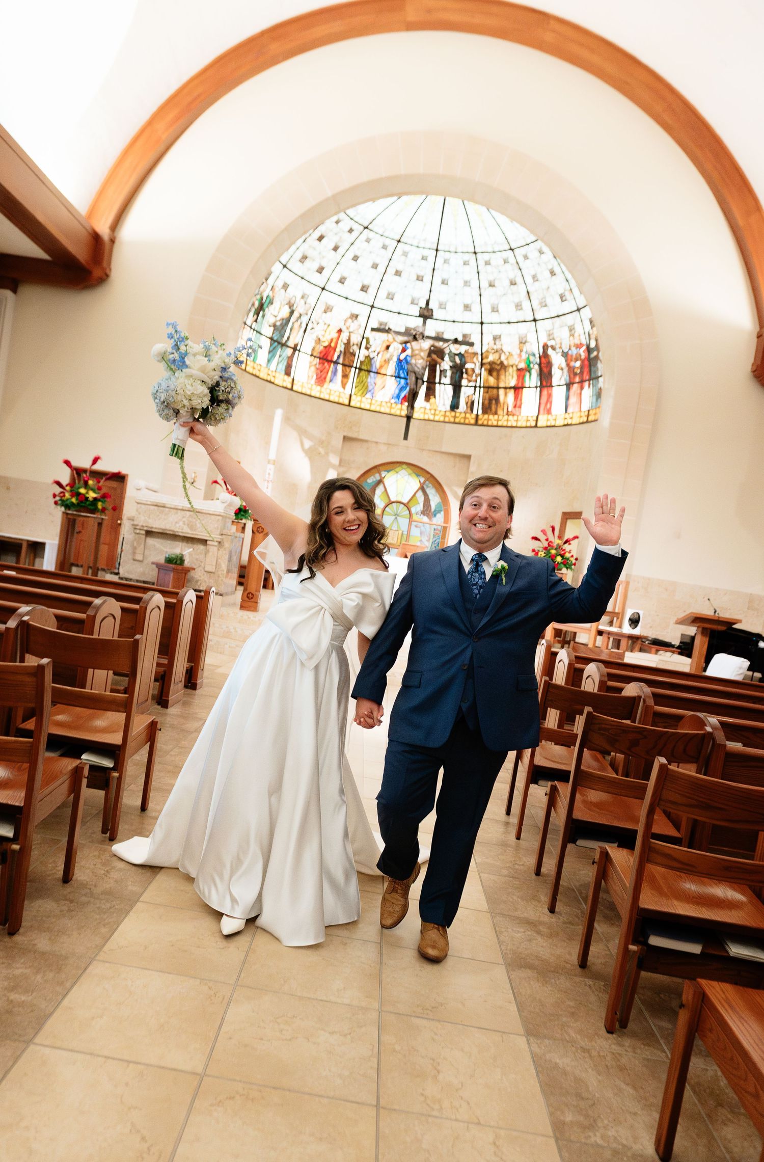 Joyful newlyweds walk down church aisle celebrating after wedding ceremony under beautiful stained glass dome.