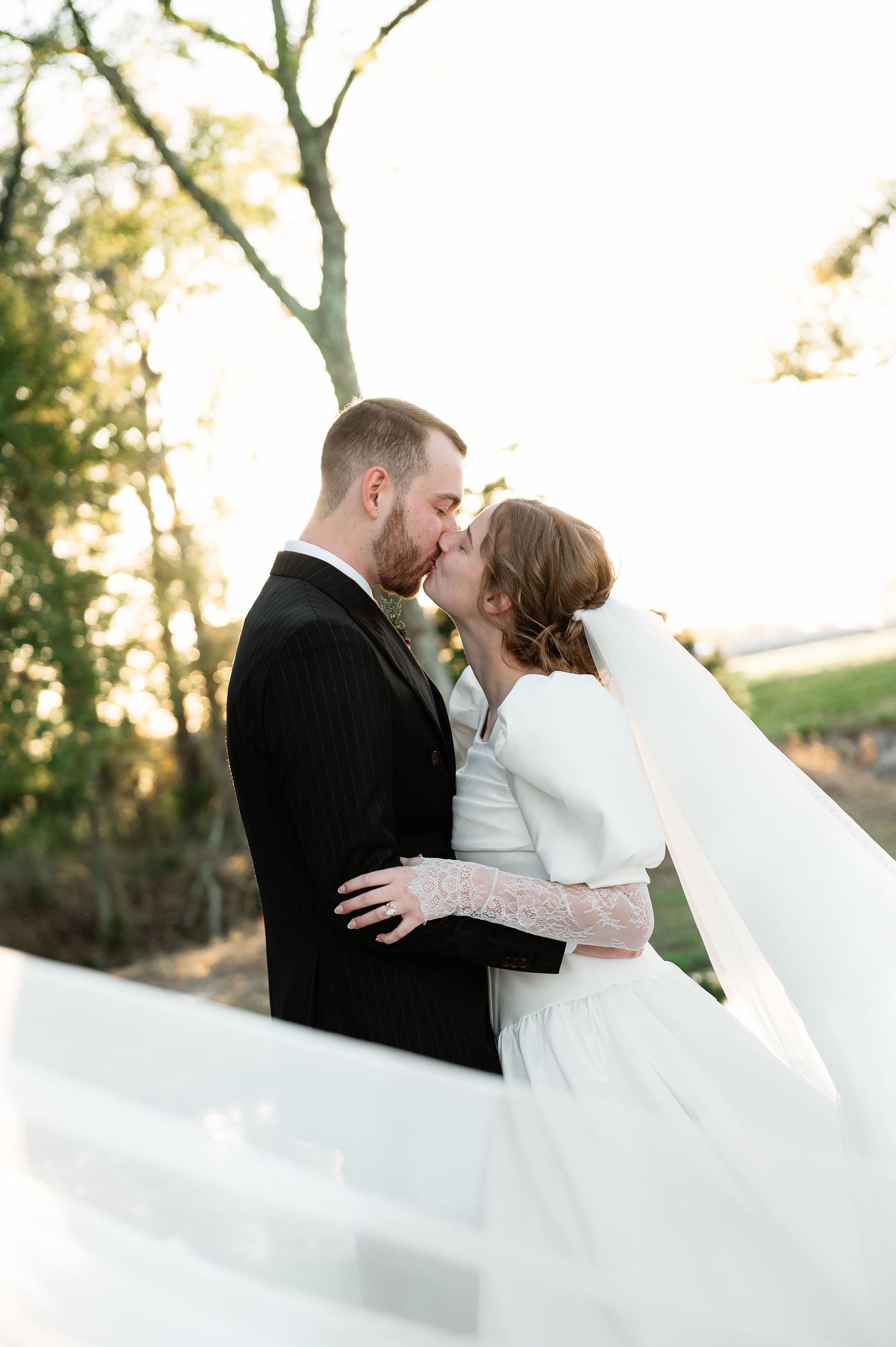 A romantic wedding couple embraces outdoors during golden hour with a flowing white dress and elegant black suit.