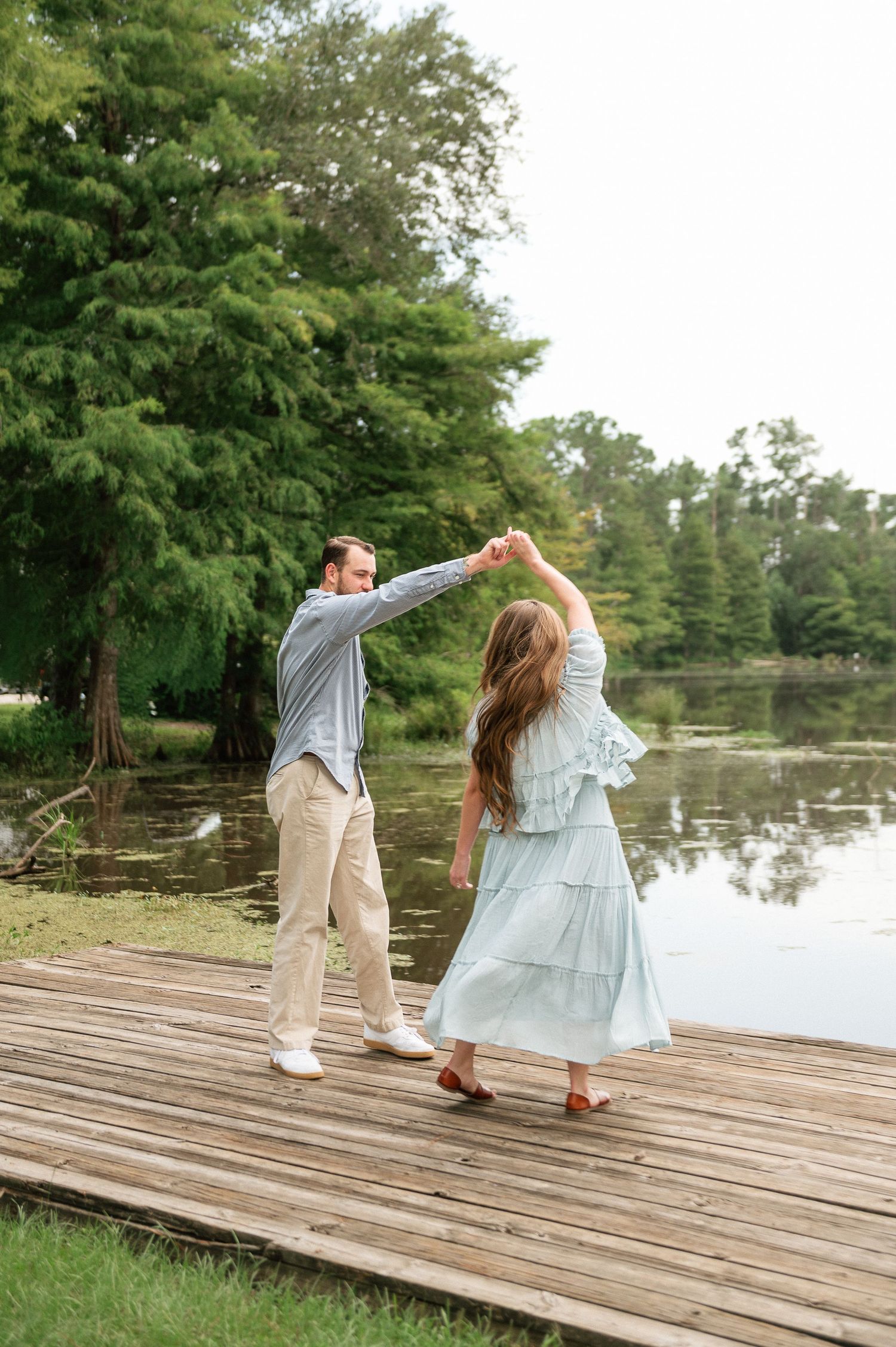 Couple dancing on a wooden dock by a serene lake surrounded by lush green trees.