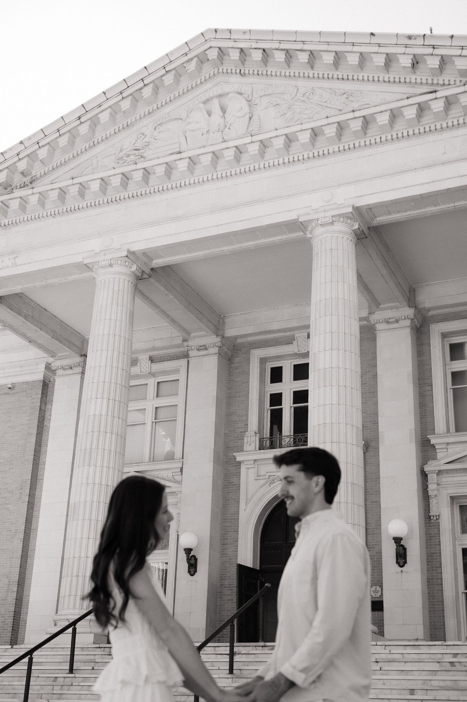 Black and white photo of a couple holding hands on steps before a grand neoclassical building with columns.
