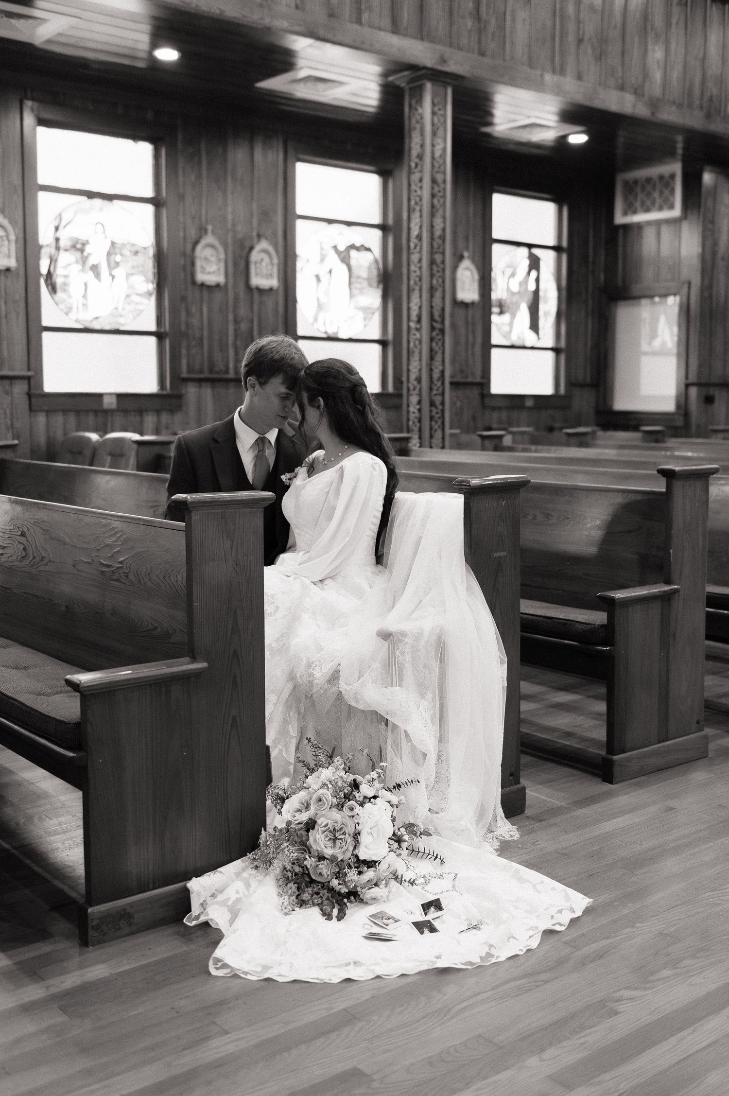 Black and white photo of a bride and groom sharing an intimate moment in a wooden church pew with a floral bouquet.