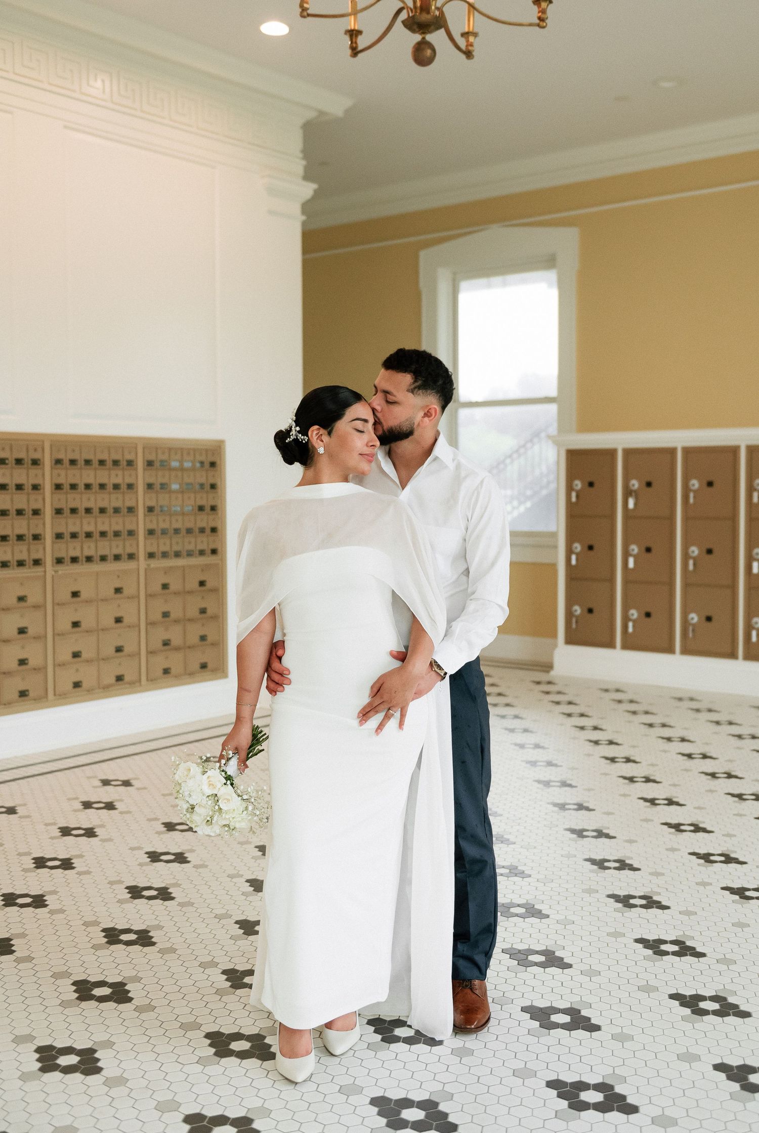 Elegant couple in wedding attire embrace in a classic interior with mosaic tile floors and vintage mailboxes.