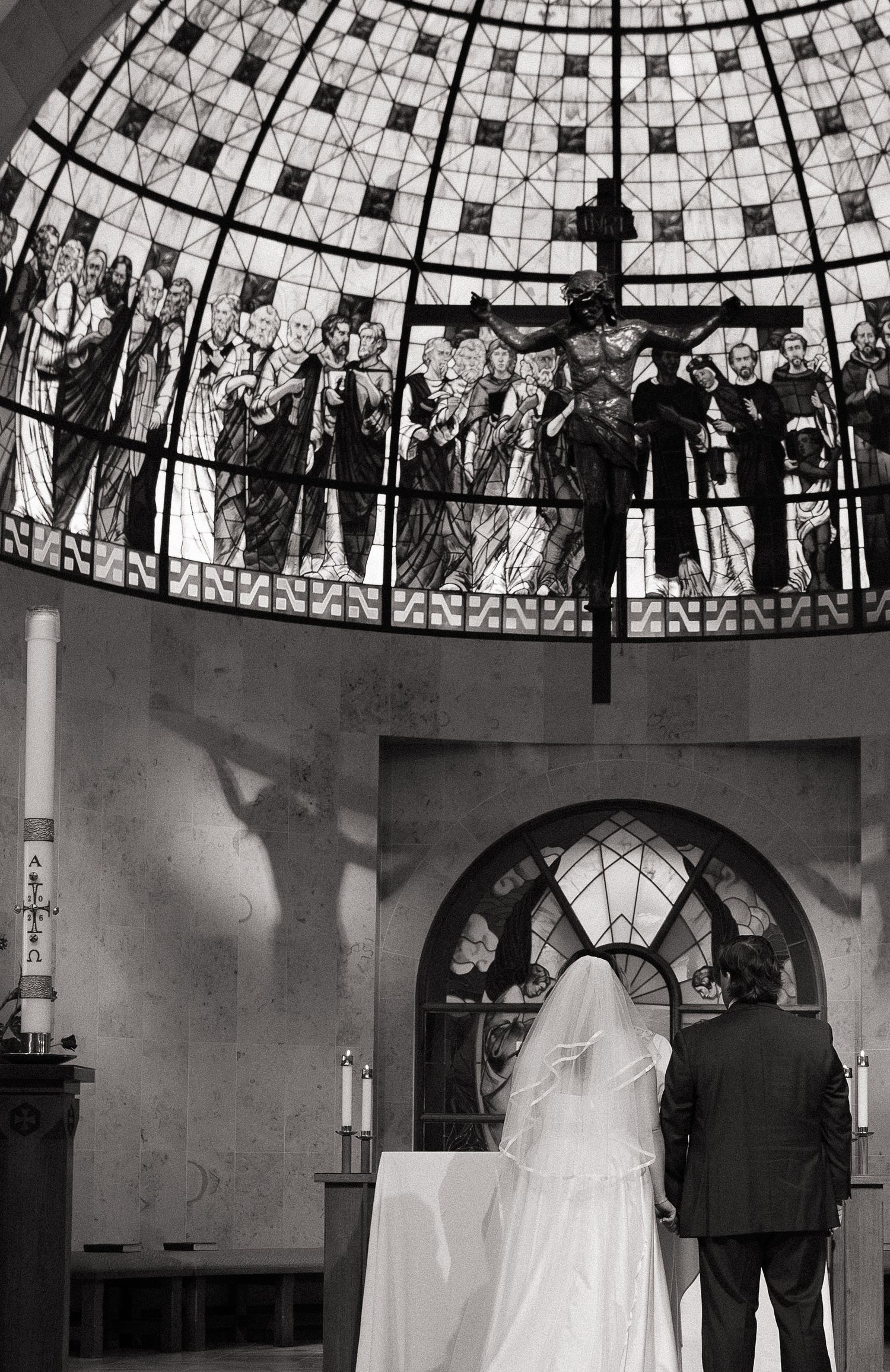 Black and white photo of a bride and groom at the altar beneath a stunning stained glass dome in a church.