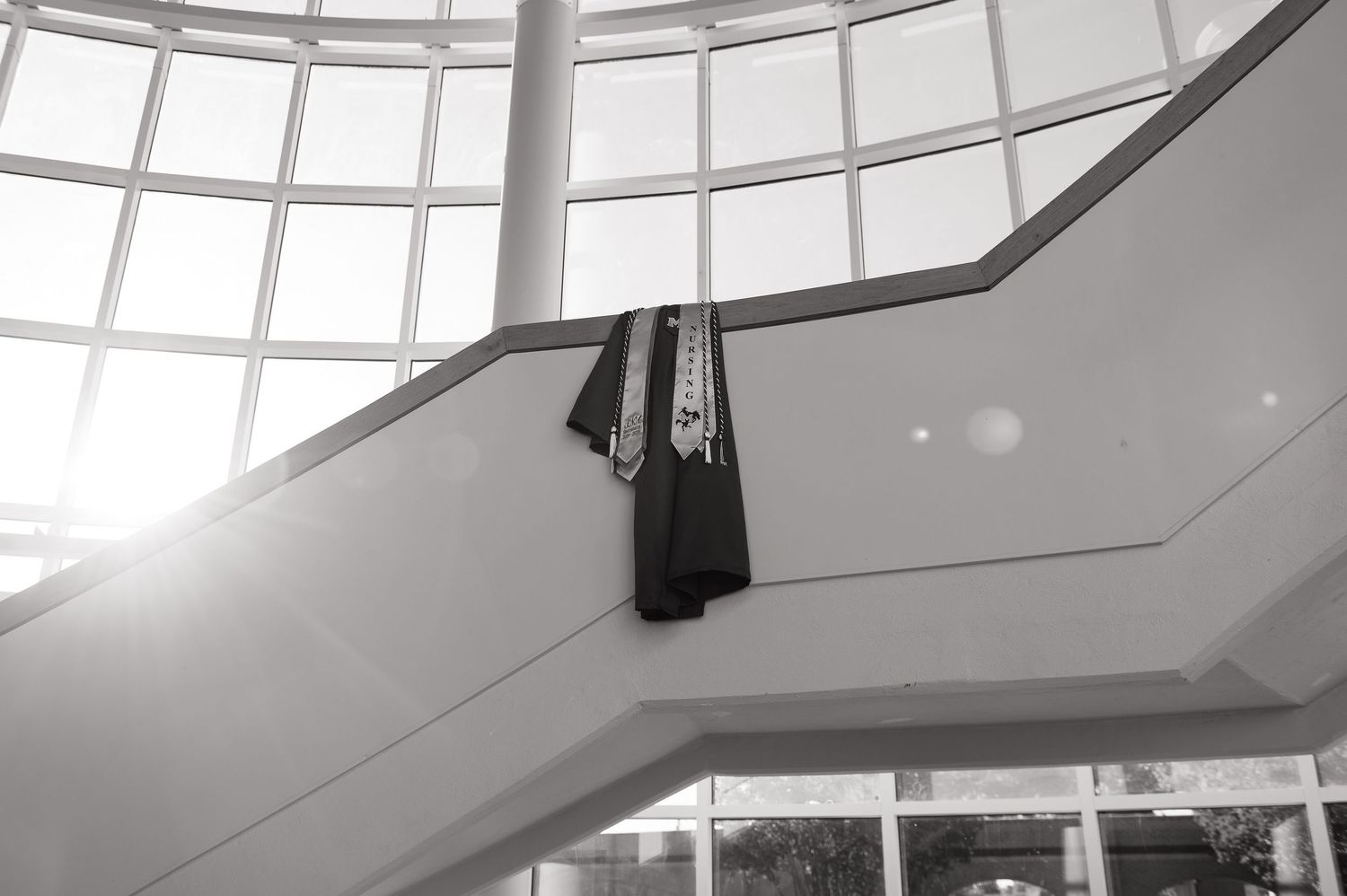 Black and white photo of a suit jacket and tie draped over a staircase railing in a modern building with large windows.