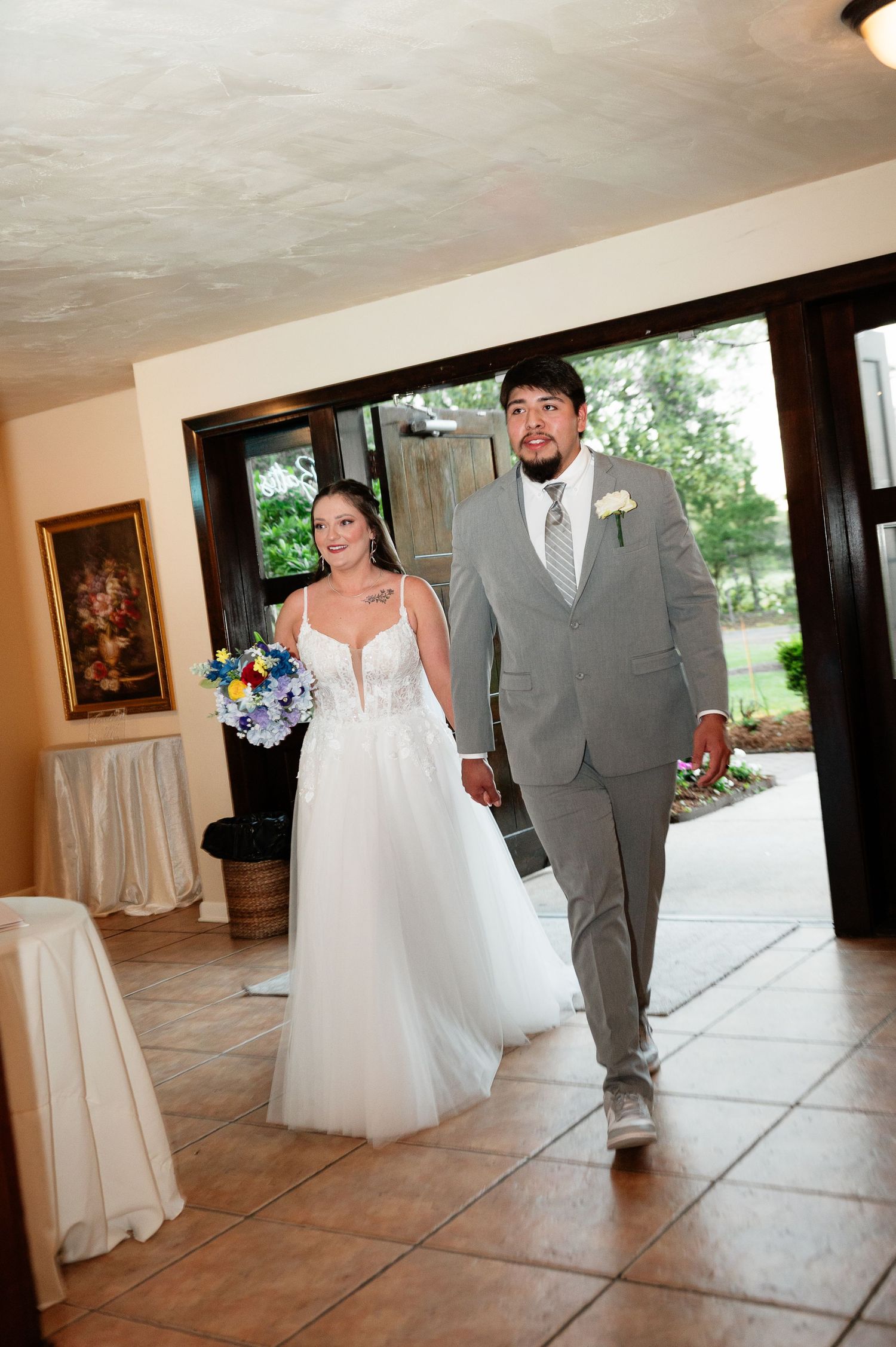 A smiling bride in white gown and groom in gray suit walk hand-in-hand into their wedding reception venue.